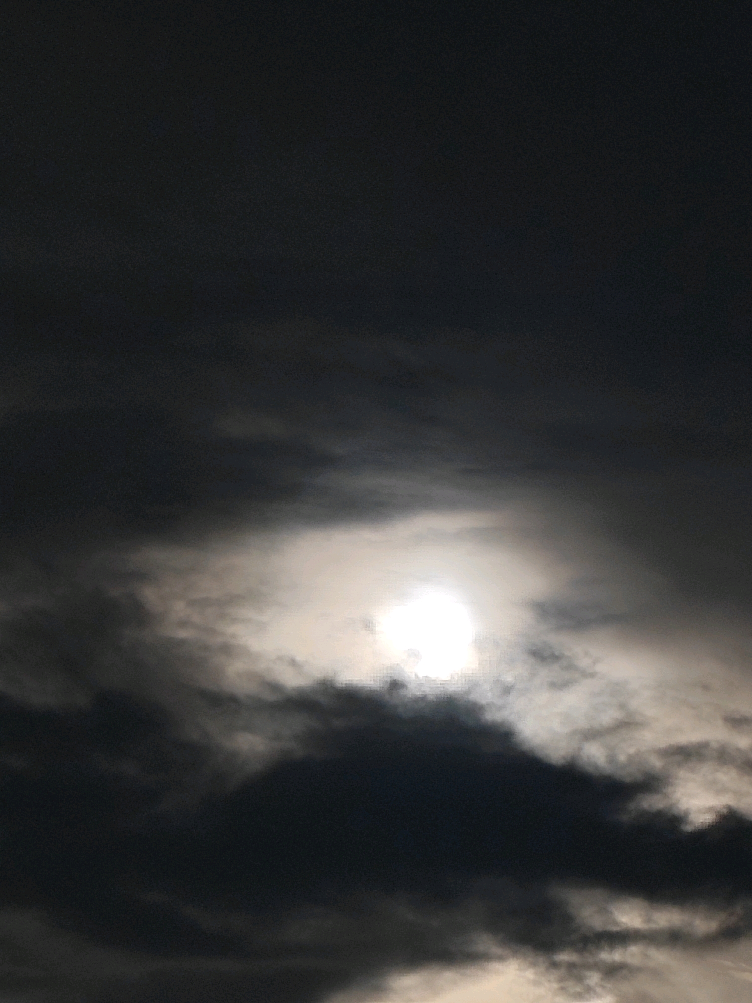 The image shows a bright, partially obscured sun high in a dark, cloudy sky. The sun is haloed by a bright white light, with dark, wispy clouds surrounding it and stretching across the frame. The lighting suggests it is either early morning or late afternoon. No specific location in Jamshedpur, India, can be identified from the image alone.