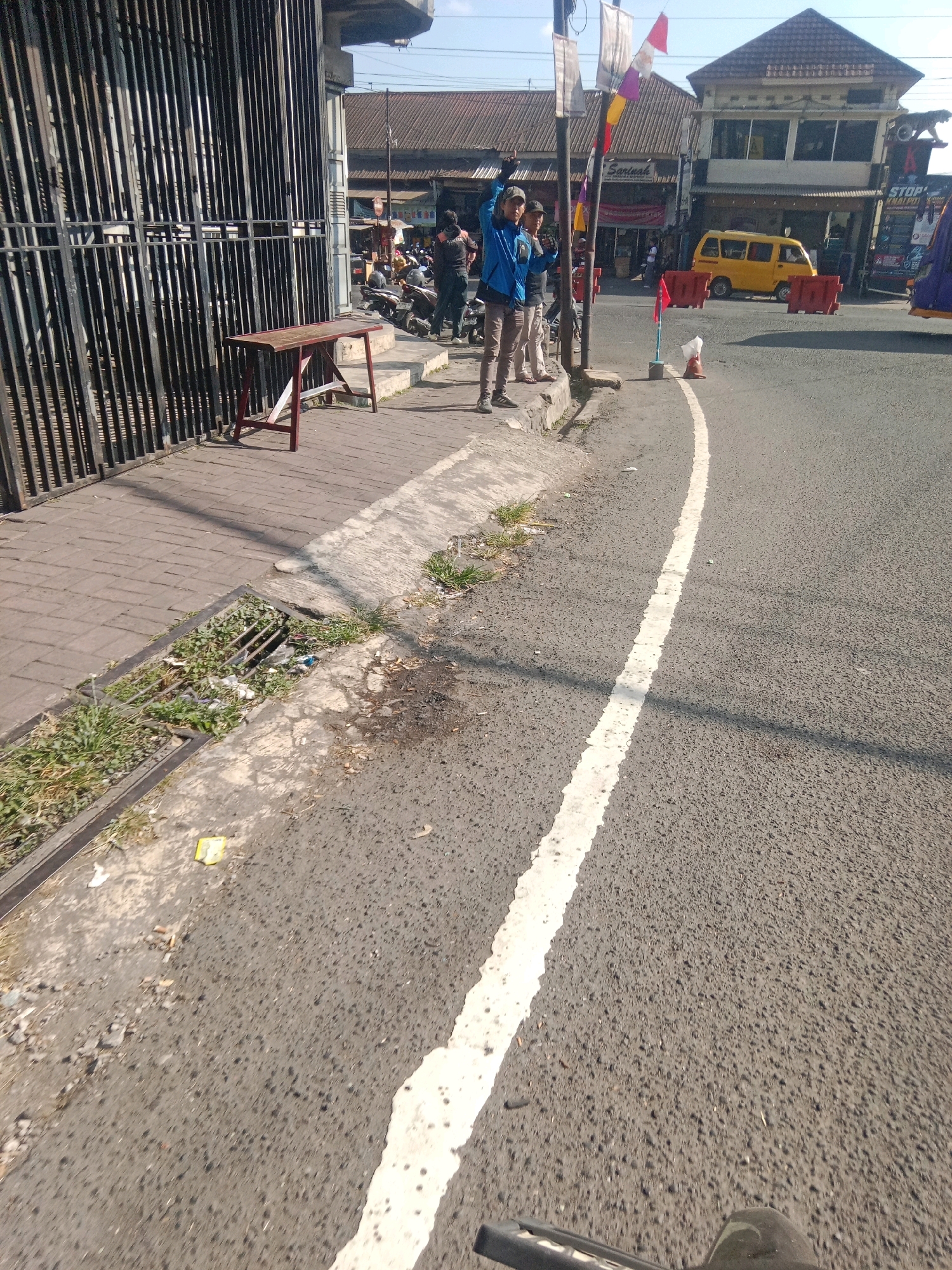 The image depicts a street scene in Kertek, Indonesia, likely near a market or commercial area. In the foreground, a paved road with a white line marking runs diagonally across the frame. A sidewalk is visible to the left, made of interlocking concrete blocks, with a metal grate draining into a ditch. Several individuals are present. Two men are standing on the sidewalk, with one in a blue jacket raising his hand. Behind them, more people are seen near parked motorcycles. A small wooden table stands on the sidewalk in front of a tall, black metal fence. In the background, commercial buildings with tiled roofs line the street. A yellow van is parked on the road, and temporary orange barriers are placed nearby. Flags and signs are also visible on poles, suggesting a public space or event. The overall environment appears to be an urban setting with moderate pedestrian and vehicle activity under a clear sky.