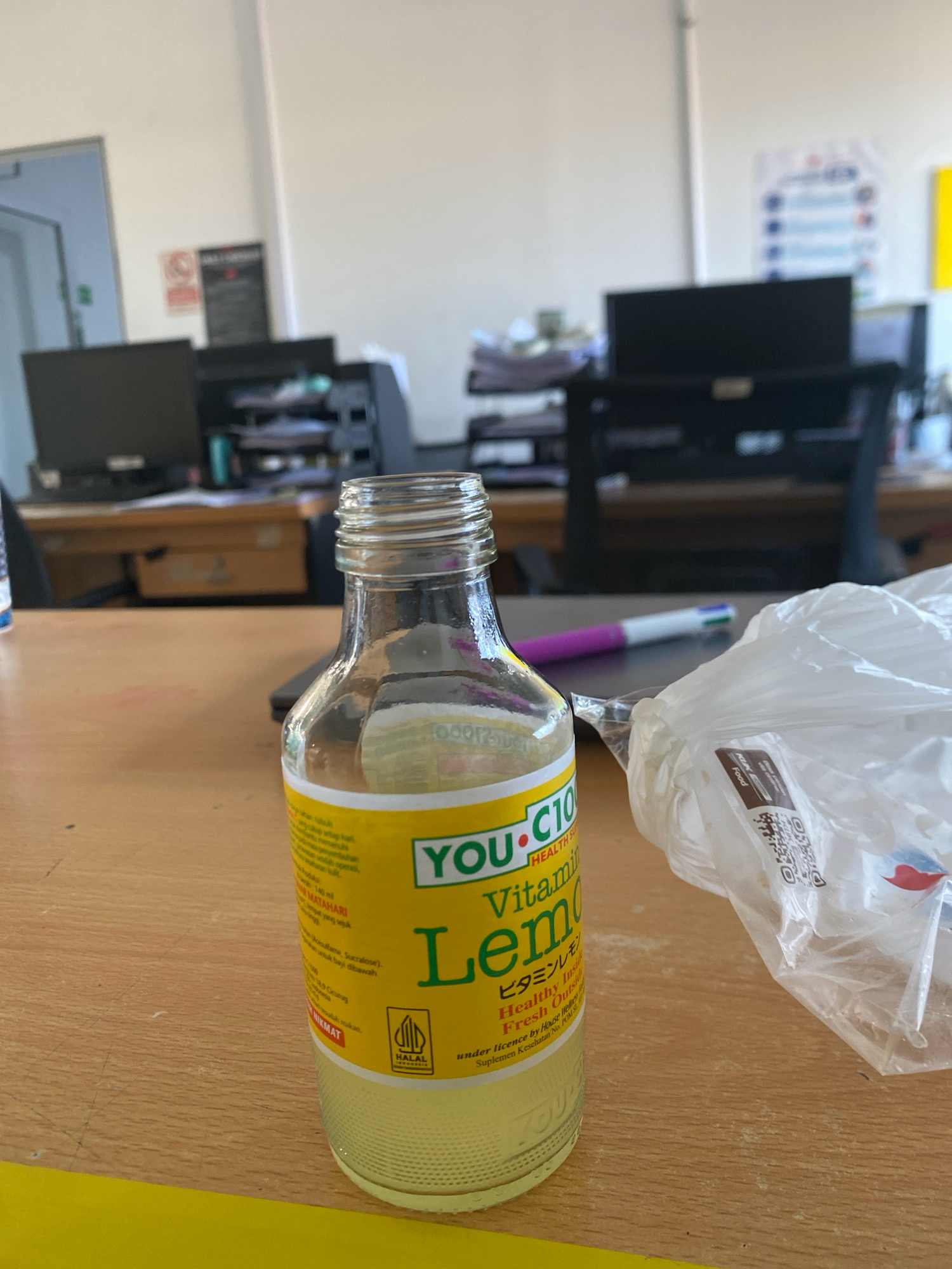 The image shows a close-up of a glass bottle of lemon-flavored drink with a yellow label, placed on a wooden desk in an office environment. The bottle is partially full of yellow liquid. The label on the bottle features text such as "YOU-C10", "Vitamin", "Lemon", "Healthy Inside Fresh Outside", and a Halal logo. To the right of the bottle is a crumpled white plastic bag, and a pen with multiple colored tips is visible in the background. Behind the desk, several computer monitors and office supplies are present, suggesting a workspace. The overall setting appears to be an indoor office. While the image does not explicitly indicate the city of Batam, Indonesia, the presence of a Halal logo on the product suggests it is likely intended for a Muslim-majority market, which aligns with Indonesia.