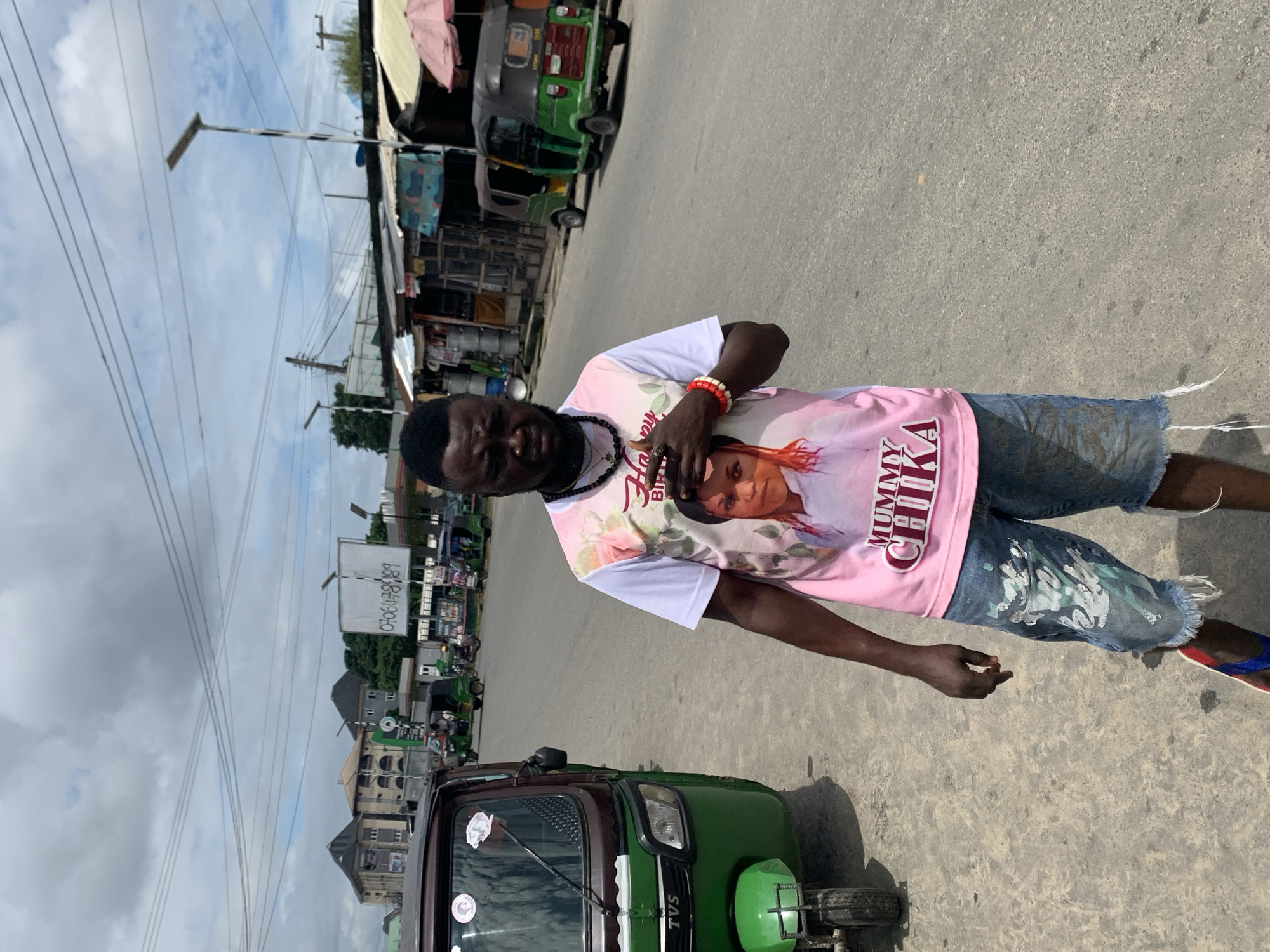 A man stands on a street in Aba, Nigeria, wearing a pink t-shirt with a picture of a woman and the words "MUMMY CHIKA" printed on it. He is also wearing denim shorts with white paint splatters and flip-flops. The man has a beaded necklace and bracelets on his wrists. He is looking at the camera with his hand on his chest.
In the background, several green and brown auto-rickshaws, also known as "Keke NAPEPs", are parked on the side of the road. There are also buildings and market stalls lining the street. The sky is cloudy. The man's stance and the t-shirt suggest he might be attending or commemorating an event related to "Mummy Chika". The presence of multiple Keke NAPEPs indicates a bustling urban environment typical of Aba, a major commercial hub in Nigeria. The general atmosphere seems to be that of a busy street scene under a cloudy sky.