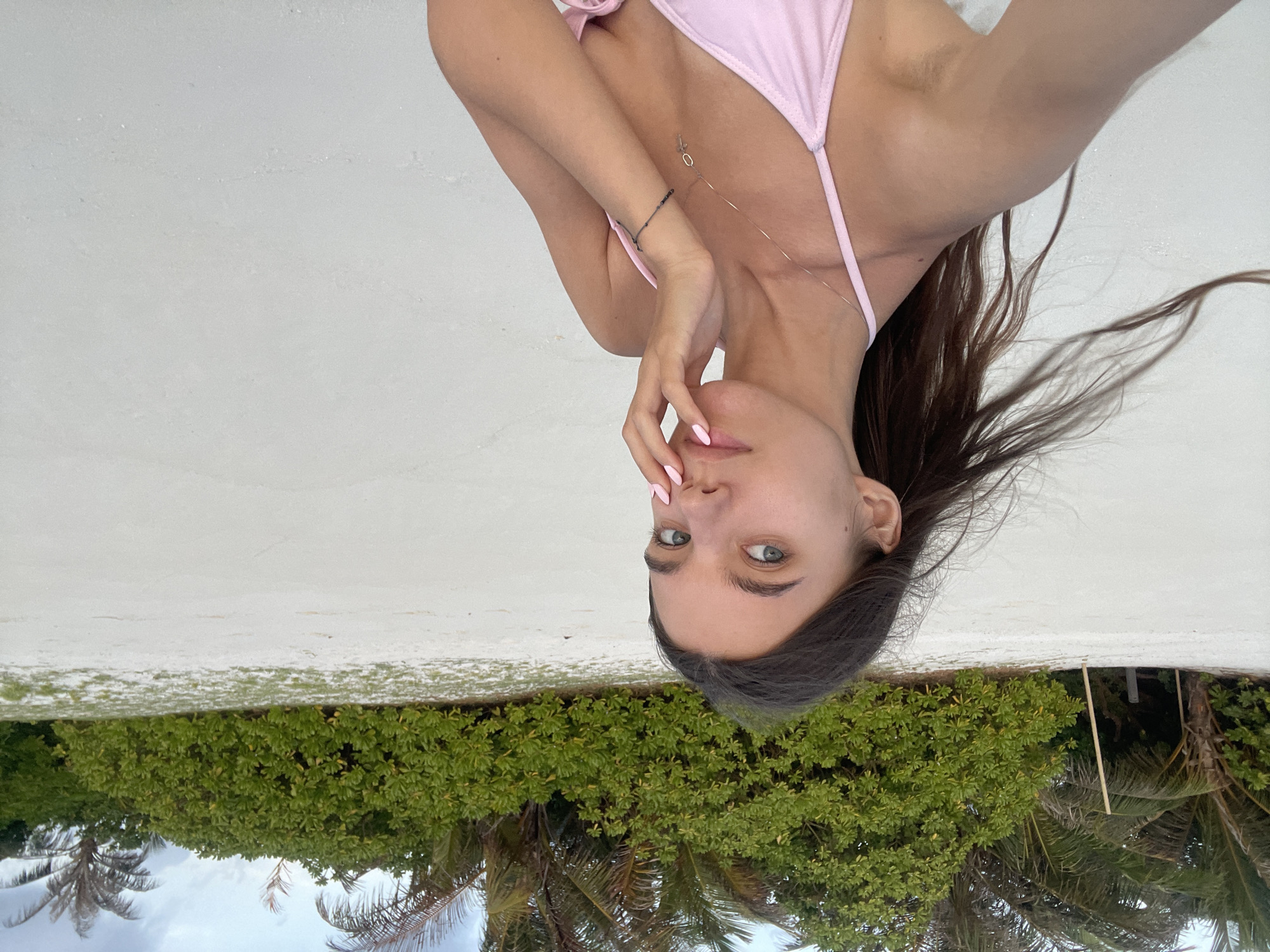 A young woman with long, dark hair is standing on a white sand beach, wearing a pink bikini top. She is looking at the camera, with her right hand touching her face. In the background, there are palm trees and lush green bushes. The sky is cloudy.