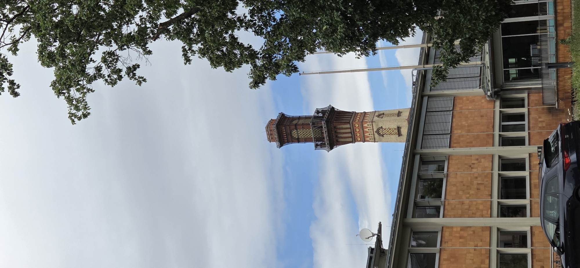 This is a photograph of the tower of the former gasworks in Karlsruhe, Germany. The tower is made of brick and has ornate yellow and red geometric patterns. It is a tall, slender structure with a distinctive crown at the top. In the foreground, there is a modern-looking building with large windows and orange tiled cladding. A dark-colored station wagon is parked in front of the building. The sky is cloudy with patches of blue. A large tree with green leaves is partially obscuring the right side of the image. The scene appears to be an exterior shot taken during the daytime.