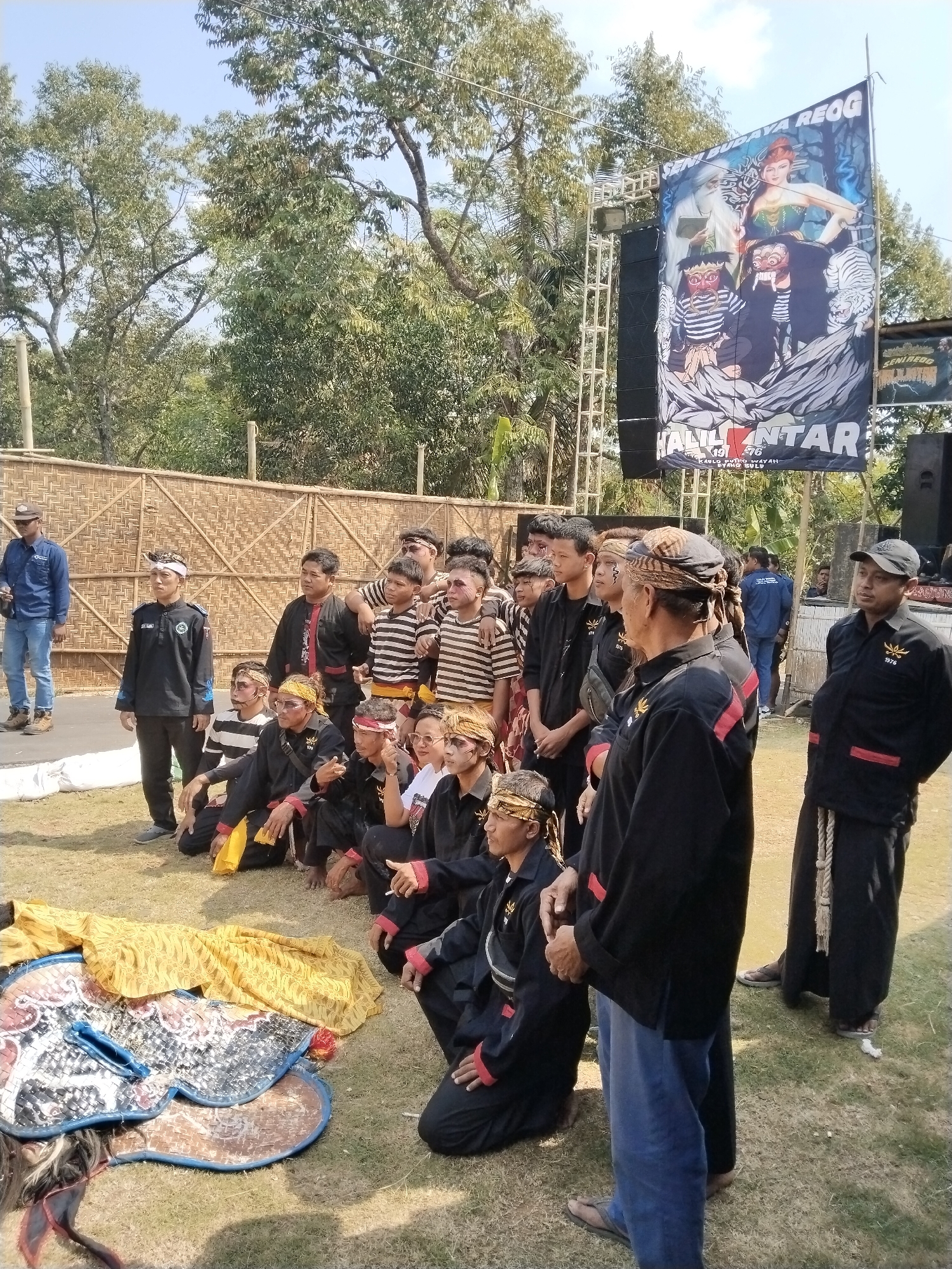 The image depicts an outdoor gathering of individuals, many dressed in traditional attire, in what appears to be a performance or cultural event. The backdrop includes a woven bamboo wall, trees, and a large banner advertising "SENI BUDAYA REOG HANTAR" with imagery of mythological figures and animals. Several individuals are posing for a photograph, some kneeling and others standing, while a few are in the background looking on. Some participants have face paint. In the foreground, two ornate masks and a yellow fabric are laid out on the grass, possibly props for the event. A man in a blue shirt and jeans stands to the left, appearing to be an observer or security personnel. The ground is covered in dry grass, suggesting an open field or park setting. The overall scene conveys a sense of community and cultural celebration. Based on the banner, this event is likely associated with the Reog Ponorogo cultural performance and may be taking place in Mojogedang, Indonesia, given the prompt's context.
