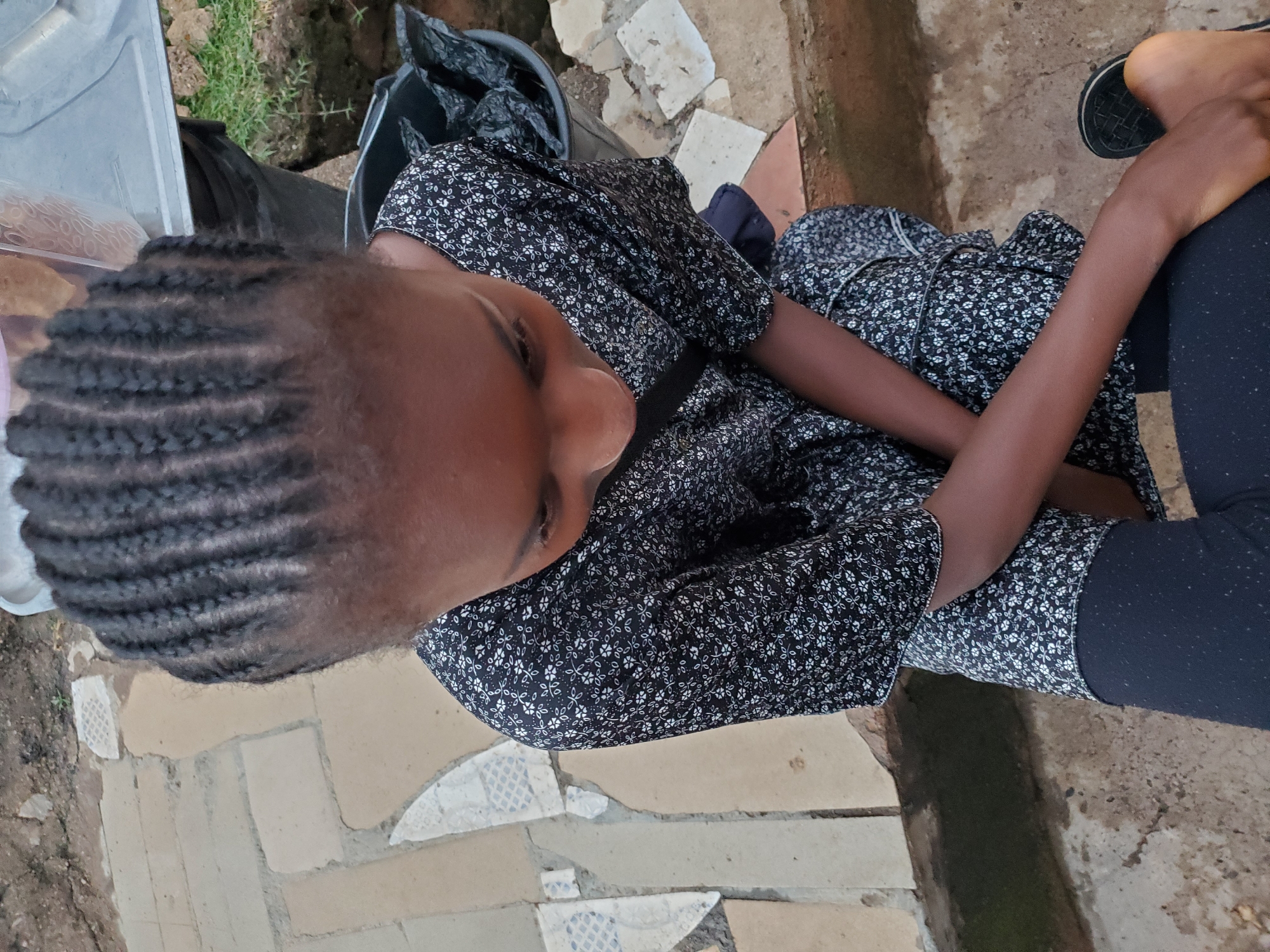 An overhead shot shows a young Black girl with braided hair looking down, seated on a concrete step. She wears a black dress with a white floral pattern and dark leggings. Her arms are crossed in her lap, and one foot is visible, wearing a black sandal. The background consists of a textured, uneven ground with scattered paving stones and some greenery. A black plastic bin filled with trash is visible to her right. The scene appears to be in an outdoor urban setting, possibly a residential area, in Jos, Nigeria.