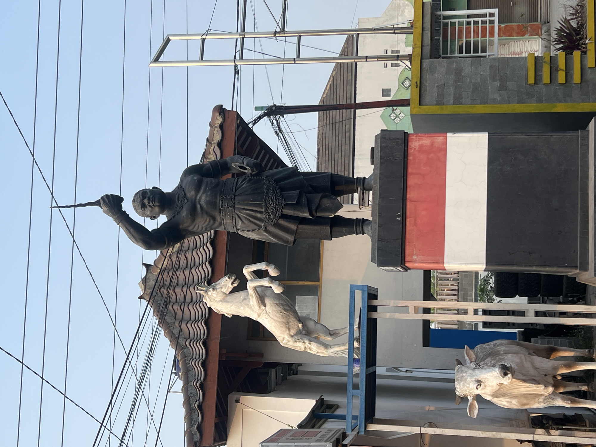 The image depicts a public monument in Tlogoadi, Indonesia, featuring a statue of a man in traditional attire, holding a spear aloft. To the left, a white horse statue is positioned on a blue platform, and a white cow statue stands to the far left. The monument's pedestal is painted with horizontal stripes of red, white, and black. The background shows buildings with tiled roofs and a network of overhead electrical wires against a clear sky.
