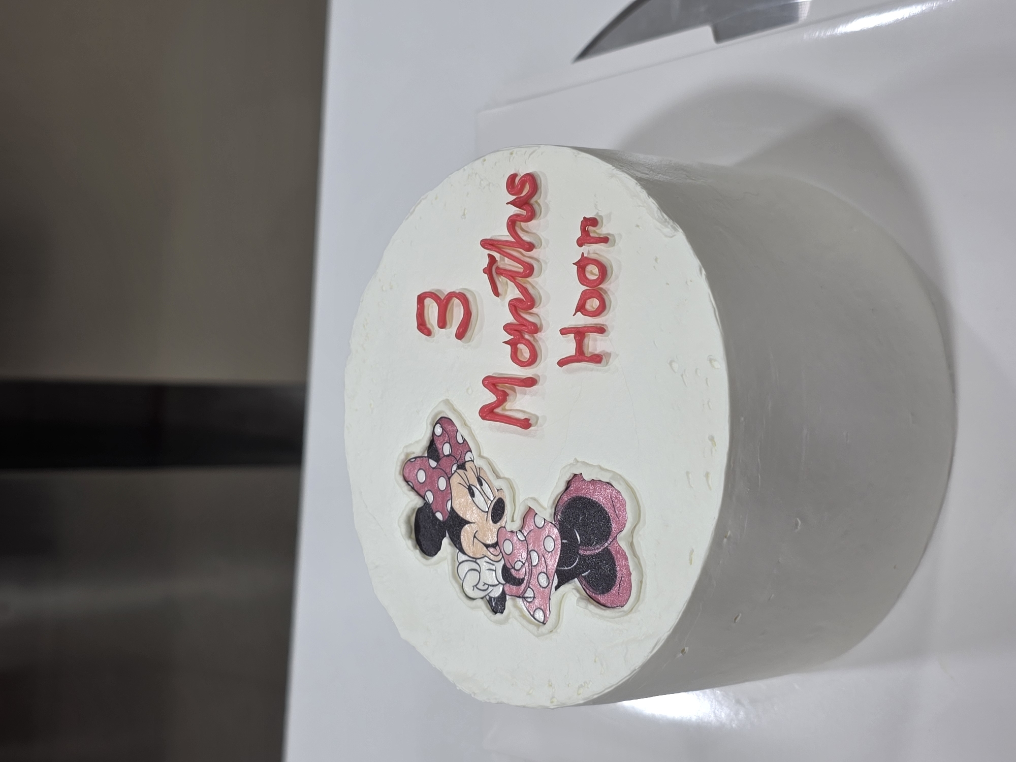 A close-up, high-angle shot shows a round, white frosted cake with a Minnie Mouse decoration and red lettering on top. The Minnie Mouse image is on the left side of the cake, showing her wearing a pink polka dot dress and bow. To the right of the image, red frosting spells out "3 Months Hoor". The cake is on a white surface, possibly a plate or cake board, with a portion of a silver knife visible on the right edge of the frame. The background is out of focus and appears to be a kitchen environment. The location of Dibba Province, Oman, is not depicted in the image.