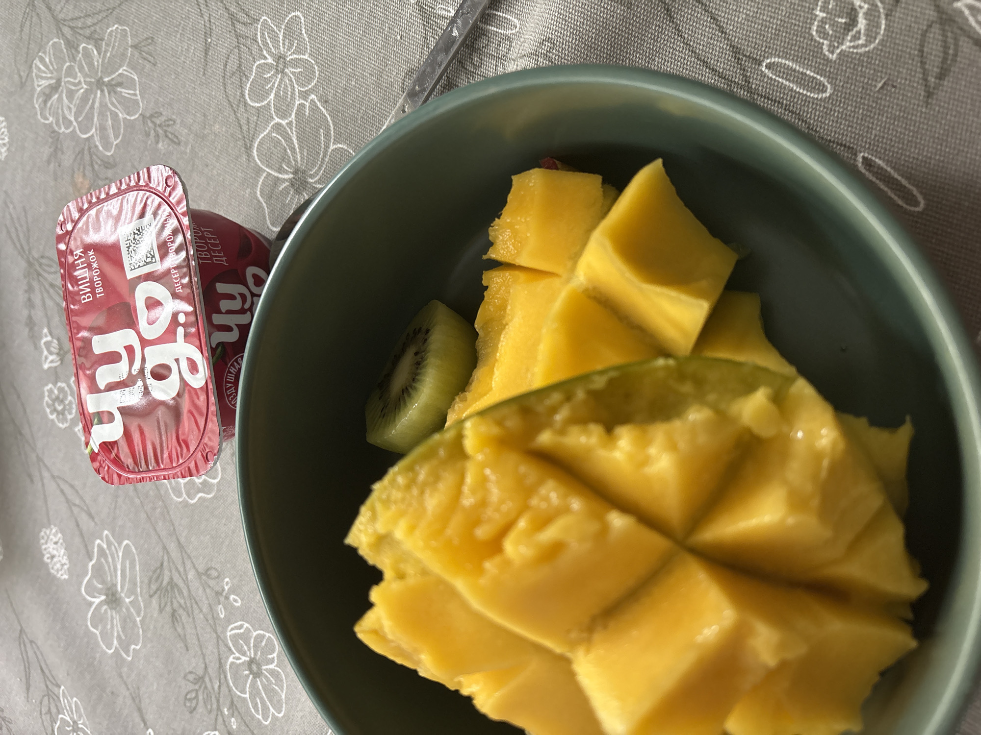 The image shows a bowl of cut mango and kiwi on a table next to a container of yogurt dessert. A spoon is near the bowl. The yogurt container has the words "ВИШНЯ ТВОРОЖОК ДЕСЕРТ" and "ЧУДО" (Cherry Curd Dessert Miracle) printed on it in Russian. The tablecloth has a floral pattern. This image was taken in a location that may be linked to Chaikovsky, Russia due to the brand of dessert.