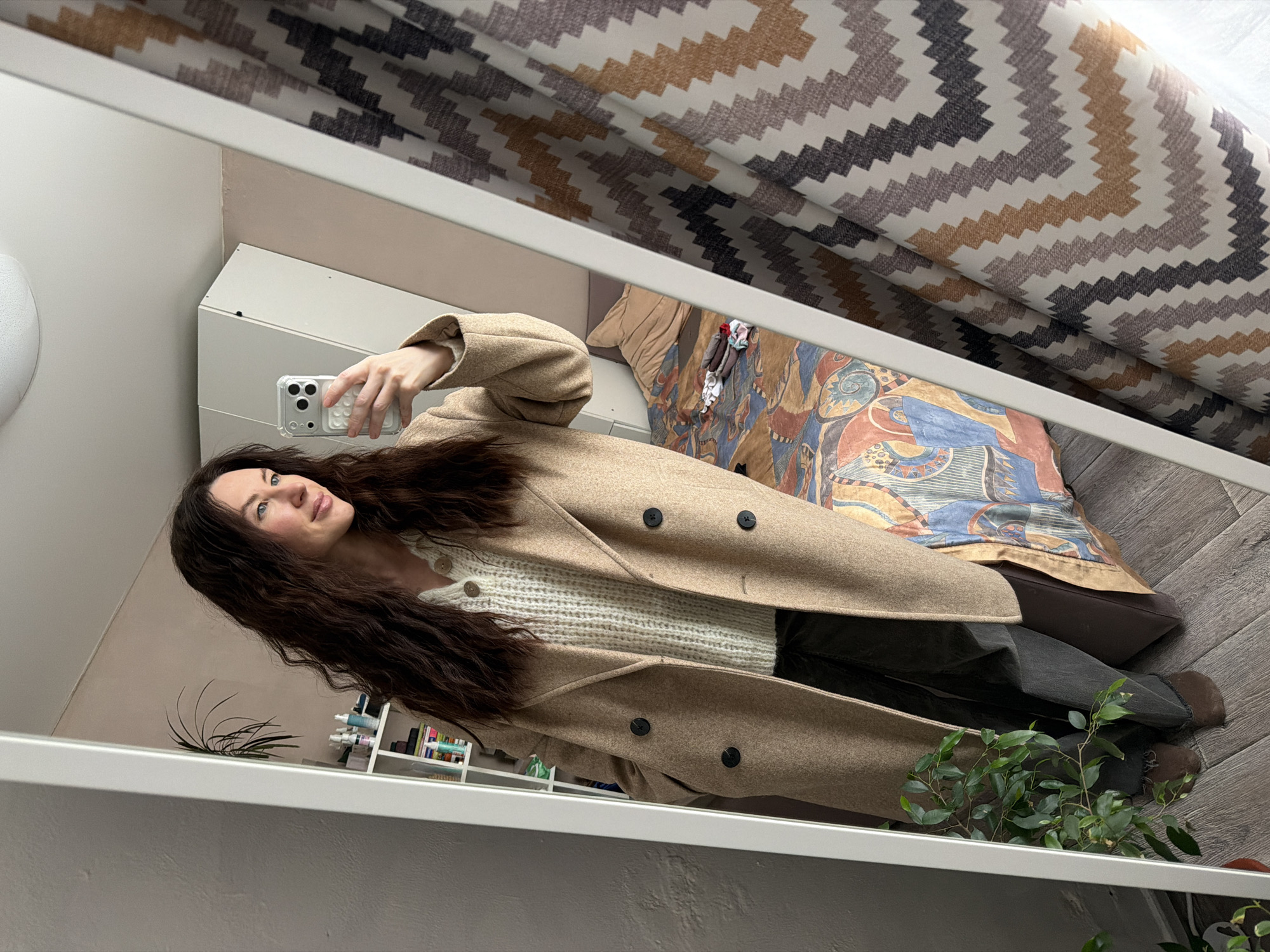 Within a domestic interior located in Perm, Russia, a female subject is depicted in a full-length mirror selfie. The woman has long, dark, wavy hair and fair complexion, with light blue eyes. She is wearing a light-colored, ribbed knit sweater with a collar and visible buttons, layered under a long, beige, double-breasted coat with dark buttons. She also wears dark trousers and dark brown flat shoes or slippers. Her right hand holds a light-colored smartphone, equipped with a clear or white textured case, positioned to capture her reflection. Her left hand is placed within the coat's pocket.
The mirrored reflection reveals details of the room. To the left, a white shelving unit holds various bottles and cosmetic items, with a green houseplant positioned below. Above this, a white ceiling fixture is visible. To the right, a bed is present, covered with a multi-colored duvet featuring abstract and geometric patterns in shades of blue, orange, and brown. A plain light brown pillow and several folded clothing items or socks rest on the bed. A white cabinet or wardrobe is partially visible above the bed. The room features a light-colored wall and a light-colored wooden or laminate floor. A curtain with a prominent zigzag pattern in white, beige, brown, and grey hues is seen on the far right.