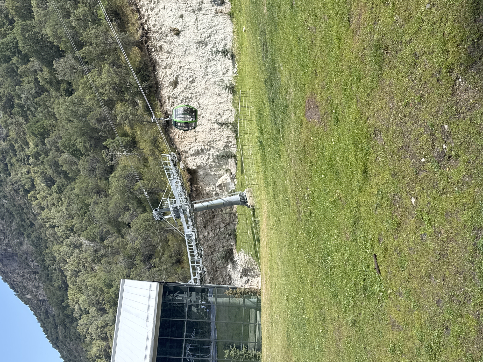 An aerial tramway gondola, which is green, is ascending a steel cable that stretches across the frame from a support tower to a building. The building, which has a metallic roof and large glass windows, is situated on the left side of the image. A grassy slope is in the foreground, and a rocky cliff face covered in trees is in the background. The sky is visible above the trees and cliff. There are no people visible in the image.