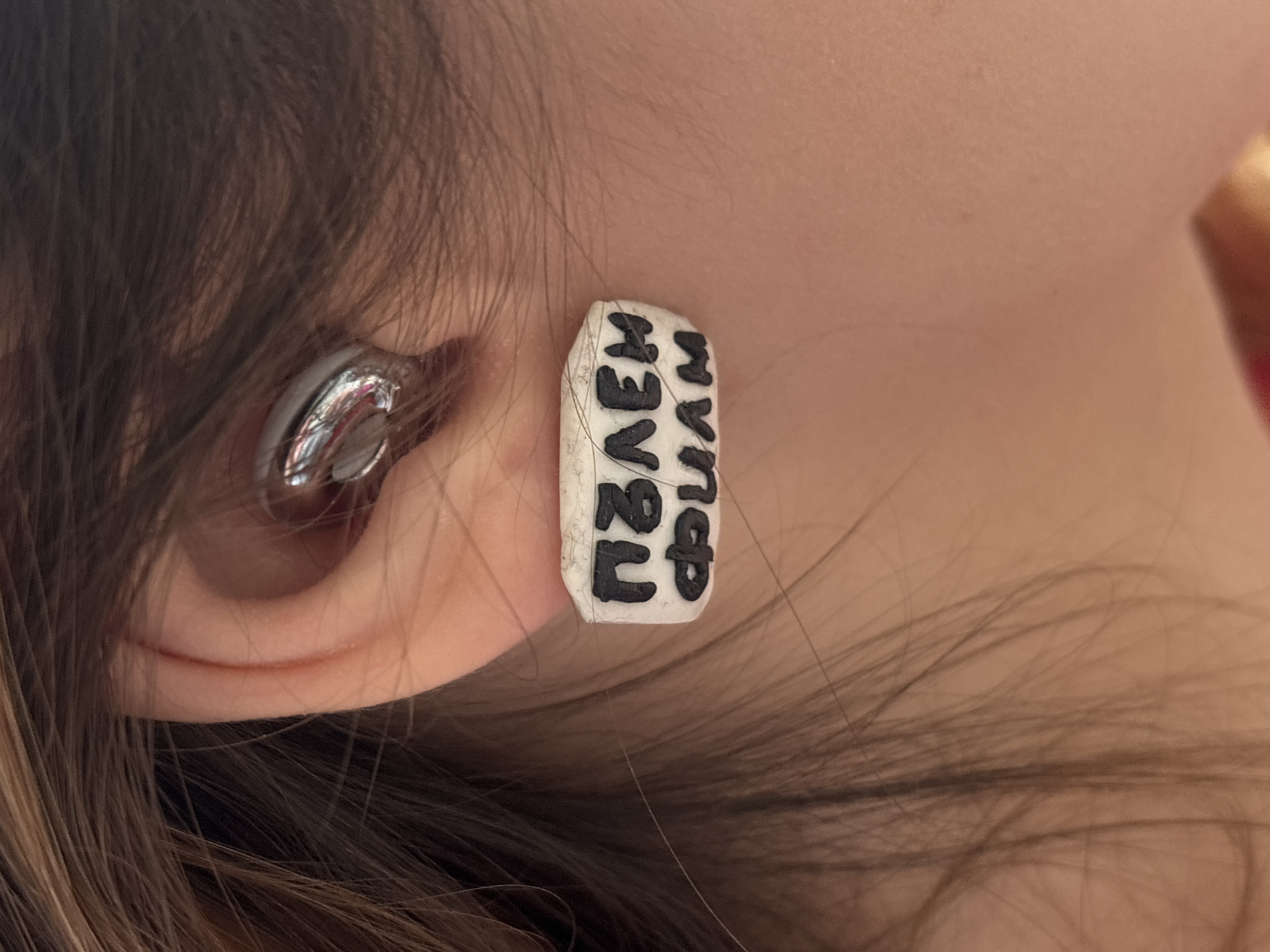 A close-up image shows a person's left ear with two earrings. The first earring is a small, silver-colored stud earring. The second earring is larger and rectangular with white and black lettering. The dark hair of the person partially obscures the view of both the ear and earrings. The lettering on the rectangular earring is not easily readable due to the angle and partial obstruction, but it appears to be written vertically. The background is out of focus. The specific location in Varna, Bulgaria, cannot be determined from the image.
