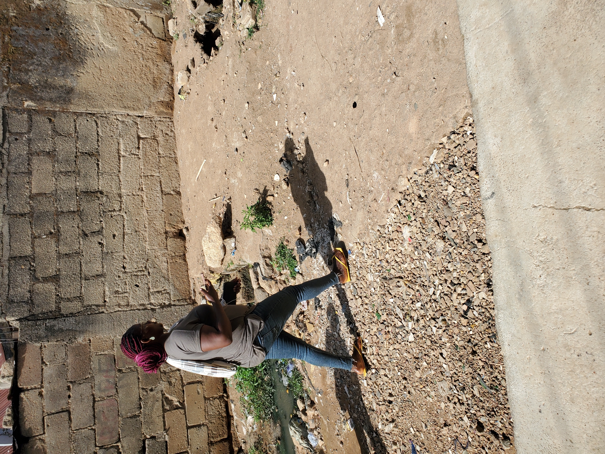 An outdoor shot shows an individual in jeans and a grey shirt, seated in what appears to be a run-down urban environment, possibly in Jos, Nigeria. The setting includes a brick wall, patches of dirt, gravel, vegetation, and some discarded waste. The individual has distinctive red hair and is holding a phone in their left hand. They are sitting on what appears to be a white object, possibly a chair or some kind of cushioning. The ground is composed of packed dirt, gravel, and interspersed debris. There is a drainage area filled with stagnant water and vegetation in the mid-ground.