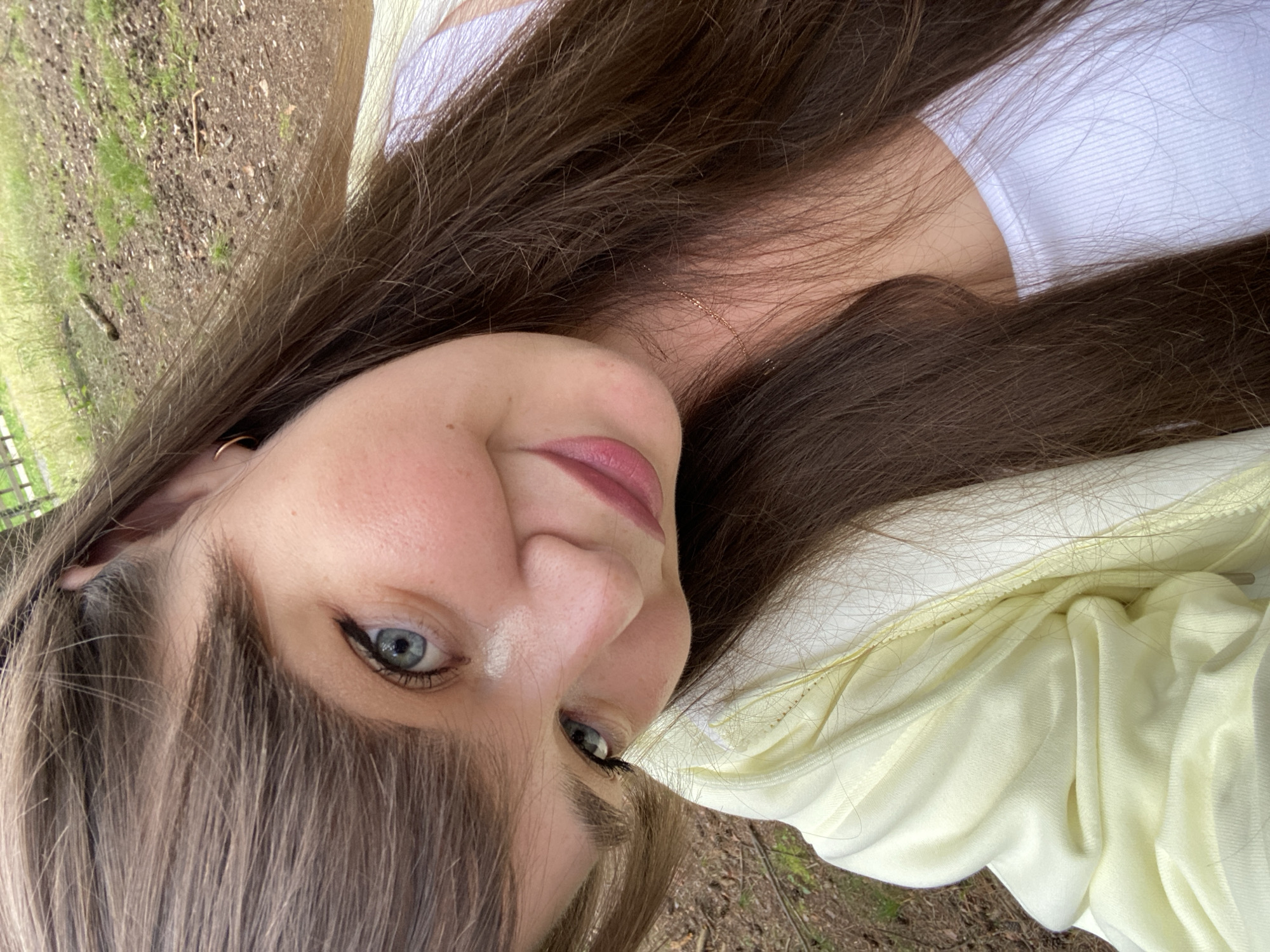 The image is a close-up selfie of a young woman with long, dark brown hair and blue eyes. She is outdoors, with a background of dry leaves, dirt, and some green grass, suggesting a park or wooded area. She is wearing a light yellow jacket over a white tank top. Her head is tilted, and she is looking at the camera with a slight smile. A delicate necklace is visible around her neck. The overall impression is a casual, outdoor portrait. There are no visible landmarks or indications of the city of Votkinsk, Russia.
