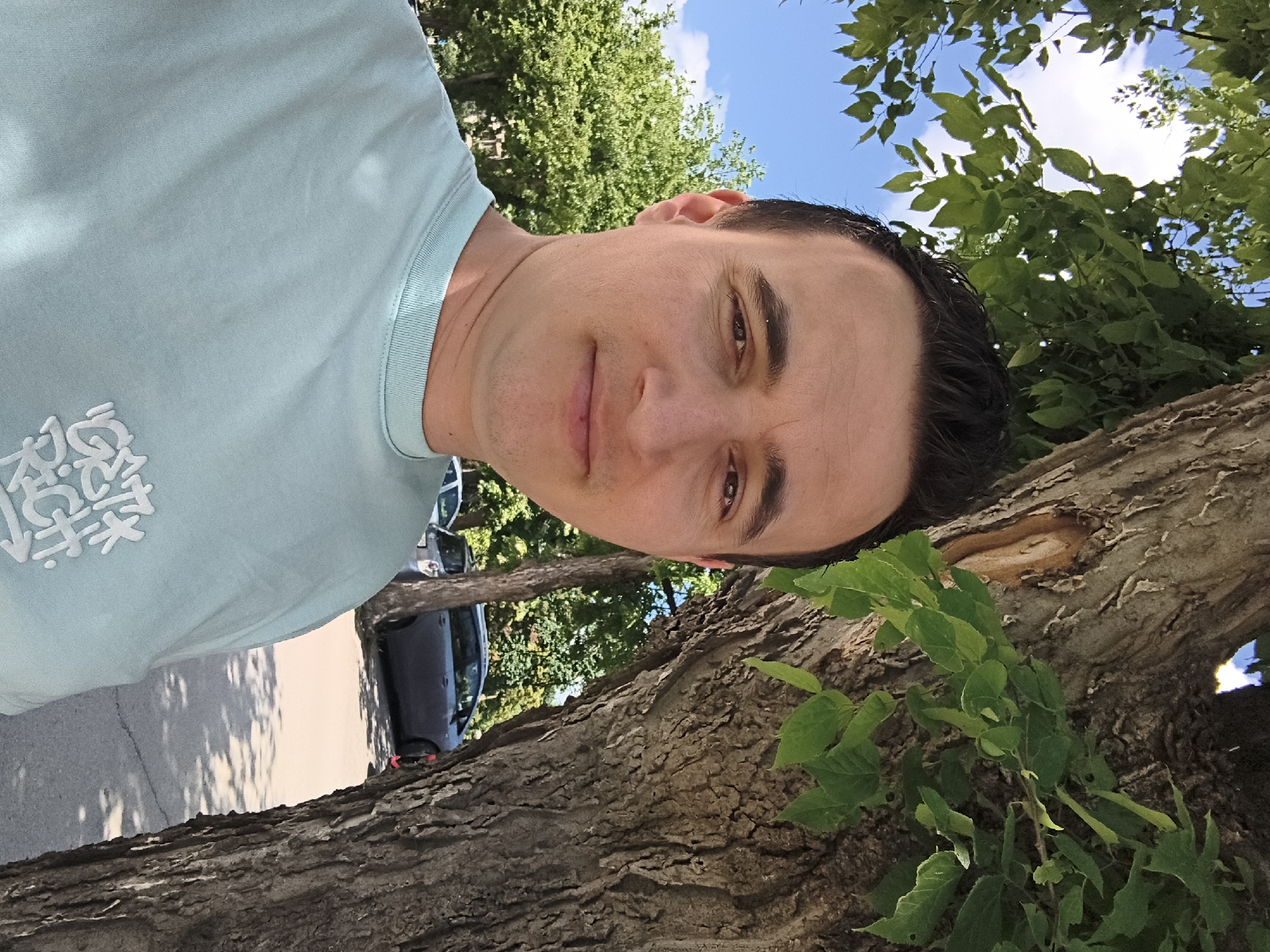 A man stands outdoors next to a tree, with cars and greenery in the background. The man is wearing a light blue t-shirt with a graphic design on it. He has dark hair and is looking at the camera. The background shows a street with parked cars, trees, and a blue sky with some clouds. The image appears to be taken in Shumen, Bulgaria.