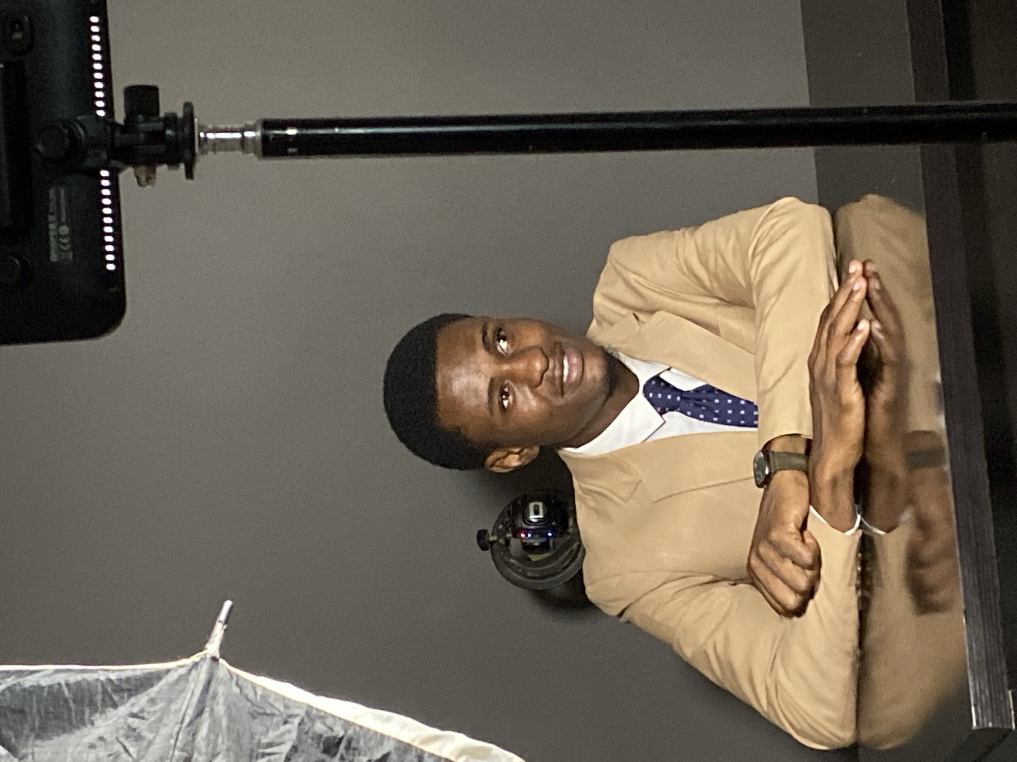 A man in a tan suit and a blue-and-white patterned tie sits in what appears to be a studio setting in Gura Top, Nigeria. There's a lighting setup visible, including a light source with LED elements, a black support pole, and what may be an umbrella or reflector. The man is wearing a watch.