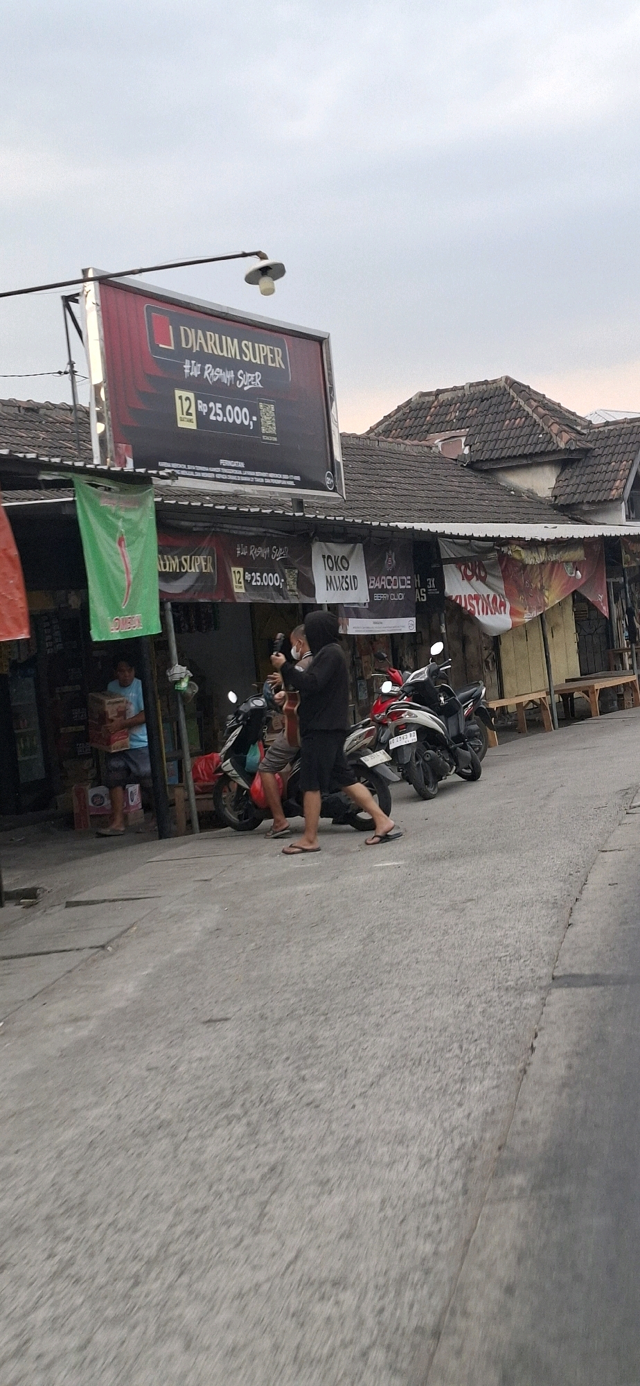 The image shows a street scene in Desa Dibal, Indonesia, with a focus on a storefront and people interacting with motorcycles. A large billboard advertising "DJARUM SUPER" is prominently displayed above a row of shops. Below the billboard, banners for various businesses like "TOKO MUSLIM" and "BARCODE" are visible.
In the foreground, three people are present. One person, wearing a blue t-shirt and shorts, stands near the entrance of a shop, holding several cardboard boxes. To their right, two individuals are standing near motorcycles. One, wearing a black hoodie and shorts, appears to be holding a phone, possibly taking a picture or video. The other, in a grey t-shirt and shorts, is also near the motorcycles. Several motorcycles are parked in front of the shops, one red and two black.
The environment suggests a commercial area, with paved road surfaces and the buildings housing the shops exhibiting traditional Indonesian architecture with tiled roofs. The lighting indicates it is likely late afternoon or early evening, with diffused sunlight and a slightly overcast sky.