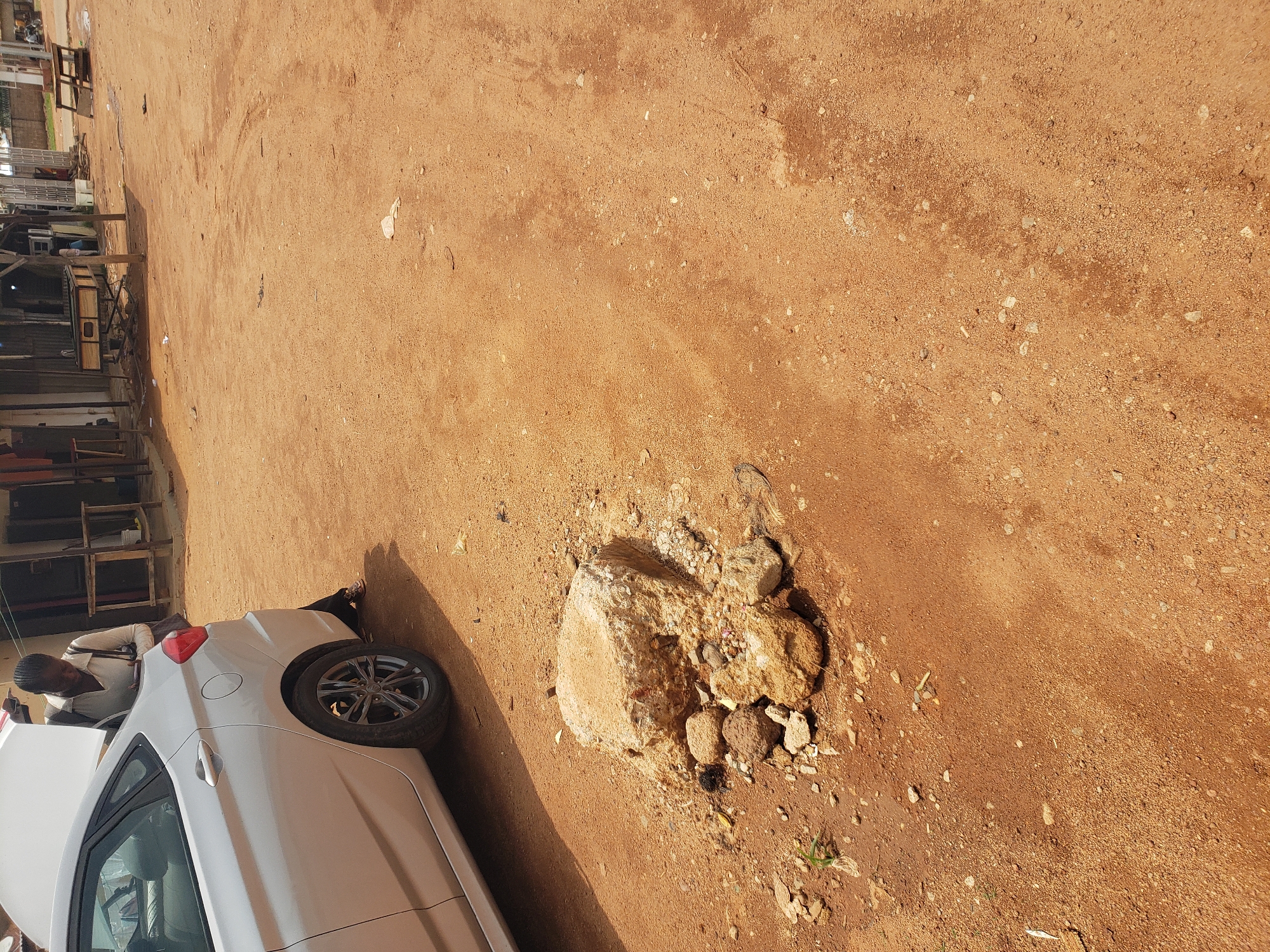 A person is standing beside a silver sedan with its trunk open, looking down into the trunk, possibly retrieving or placing an item. The car is parked on a dirt road in what appears to be a market or roadside stall area in Bukuru, Nigeria. The ground is a reddish-brown dirt, with scattered rocks and debris. In the background, there are several makeshift structures, possibly shops or stalls, constructed from corrugated metal and wood. The overall scene suggests a busy, outdoor commercial environment.