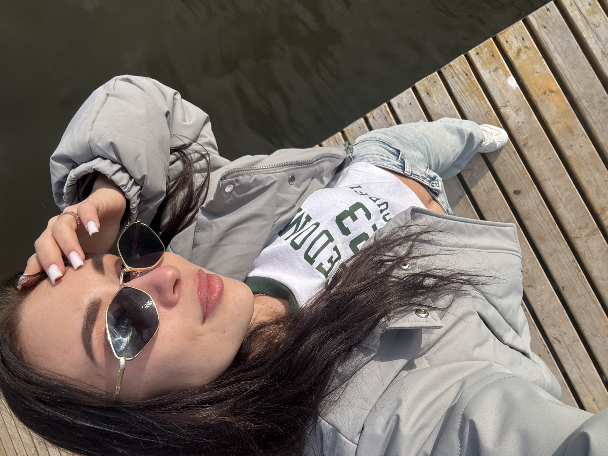 A young woman is lying down outdoors on a sunny day, captured in an overhead shot. She is positioned on a wooden pier or dock, with a dark body of water visible to her left.
**Subjects:** The primary subject is a woman with long, dark hair, wearing large, dark sunglasses with uniquely shaped lenses. She is dressed in a light grey puffer jacket, a white t-shirt with green text, and light blue jeans. Her feet, clad in white sneakers, are visible towards the bottom right. Her hands have manicured white nails and she wears a simple ring on one finger.
**Activity:** She appears to be posing for a photograph, lying in a relaxed manner with her head slightly turned towards the camera. One hand is gently placed near her temple, touching her hair or sunglasses.
**Notable details:** The scene is brightly lit by natural sunlight, casting distinct shadows and highlighting the texture of the wooden planks and the ripples on the water's surface, indicating daytime with clear weather. The wooden dock planks are weathered, showing variations in color and some light scuffs. The water looks calm and reflects the light slightly. Her expression, partially hidden by sunglasses, seems relaxed.
**Visible text:** On her white t-shirt, green text is visible, partially upside down, reading "FREEDOM" and possibly other words or numbers, though "FREEDOM" is the most discernible part.
**Setting:** The location is an outdoor setting next to a body of water, likely a lake or river, in Фоки, Russia.
