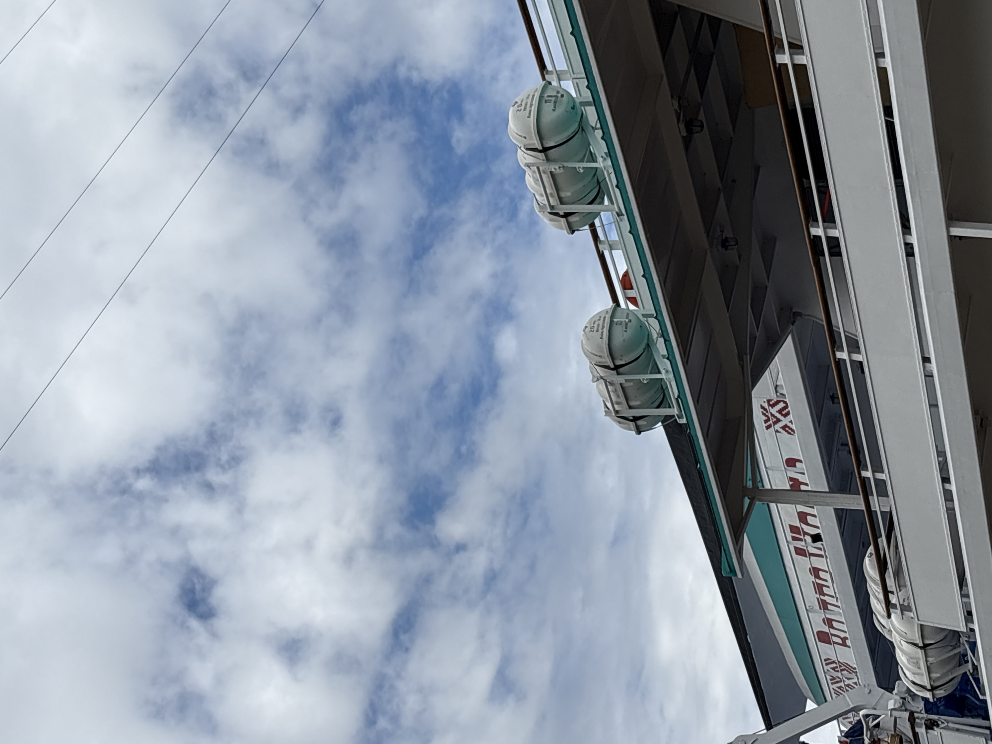 A close-up, angled view shows the upper decks of a cruise ship against a cloudy sky with visible power lines stretching across the frame. On the top deck, two rows of white, cylindrical life rafts are secured to a railing. The lower deck features the side of the ship with a decorative band of red lettering that appears to spell out "BATRANA BAMA" and a turquoise stripe. The railing on this deck is a dark brown, and the ship's structure is white. A dark shadow partially obscures the lower decks. The scene is likely at a port in Saint Petersburg, Russia, given the presence of cruise ships and typical infrastructure.