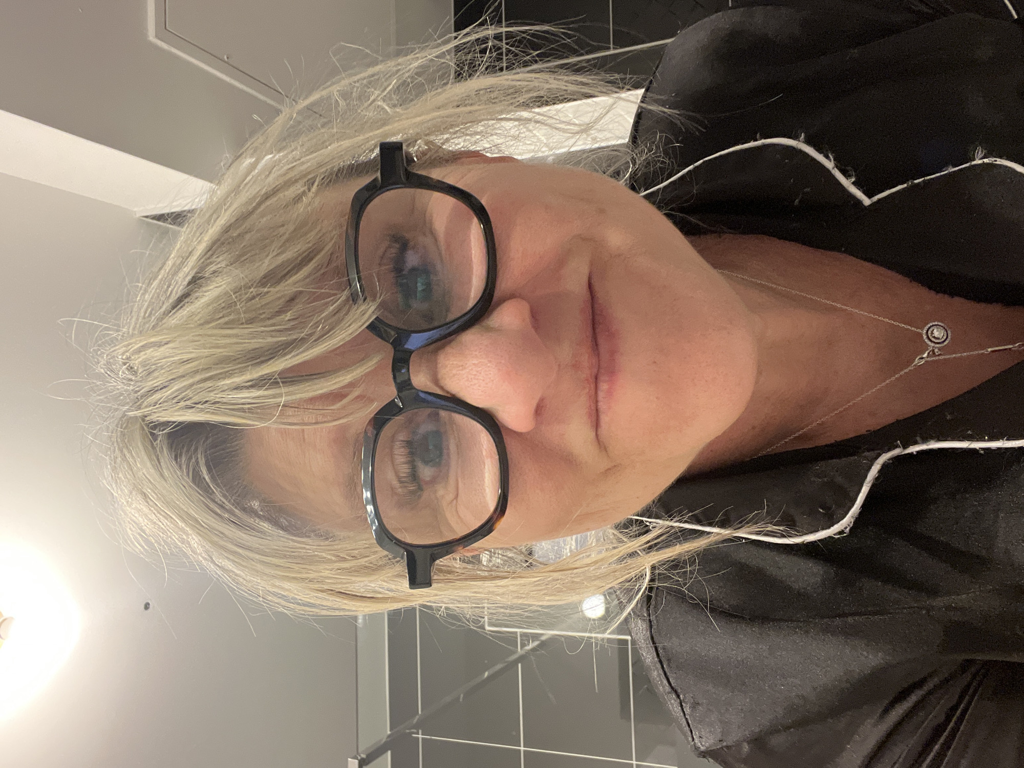 A close-up, tilted shot shows a middle-aged woman with blonde hair wearing glasses and a black top with white piping. She is wearing a delicate silver necklace. The background includes light grey and black tiles, and a section of a light grey wall. The location is not identifiable from the image.