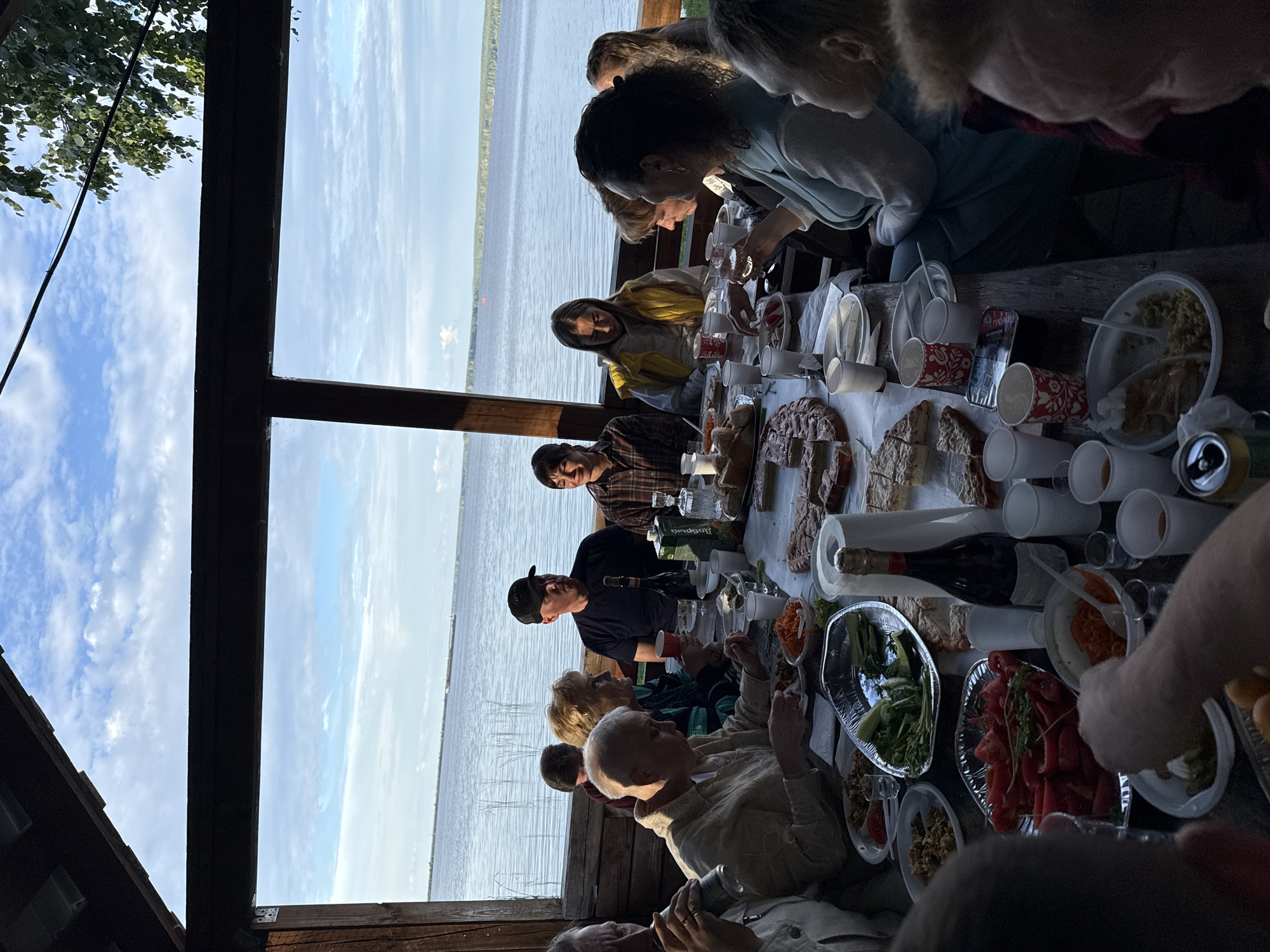 In an outdoor pavilion with a wood-paneled ceiling and open sides offering a view of a wide body of water and a cloudy sky, a group of approximately twelve people are gathered around a long wooden table laden with food and drinks. The people, of various ages and genders, are seated and interacting with each other and the food. Some are eating, others are talking, and some are looking out at the water.
On the table, there are platters of food including sliced bread, what appears to be a large pie or cake, a salad with greens and what looks like tomatoes and cucumbers, bowls of shredded carrots, and other dishes that are partially obscured. Drinks include bottles of wine, a carton of juice, and various cups and glasses. A can of soda is visible on the table.
The environment is natural, with tall reeds visible in the water near the shore and trees in the distance. The overall scene depicts a relaxed social gathering, possibly a picnic or meal shared outdoors. The lighting suggests it is late afternoon or early evening.