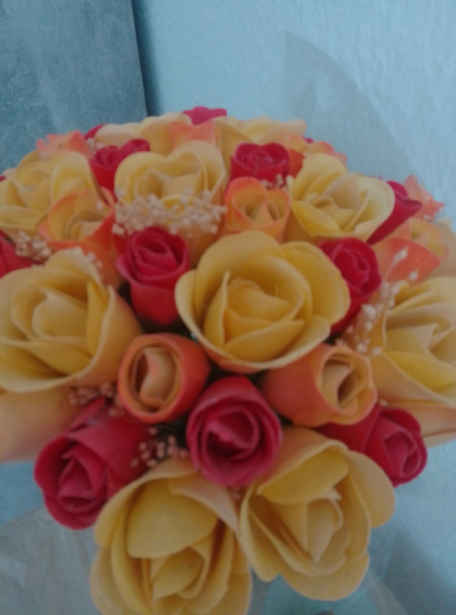 The image shows a close-up view of a bouquet of artificial roses. The roses are a mix of yellow, red, and peach colors, with some small white embellishments scattered amongst them. The background is a blurred blue surface, possibly fabric or a painted wall. The lighting is soft, and the image is slightly out of focus, giving it a dreamy or artistic quality. There are no people or specific landmarks visible in the image to indicate a location in Wedel, Germany.