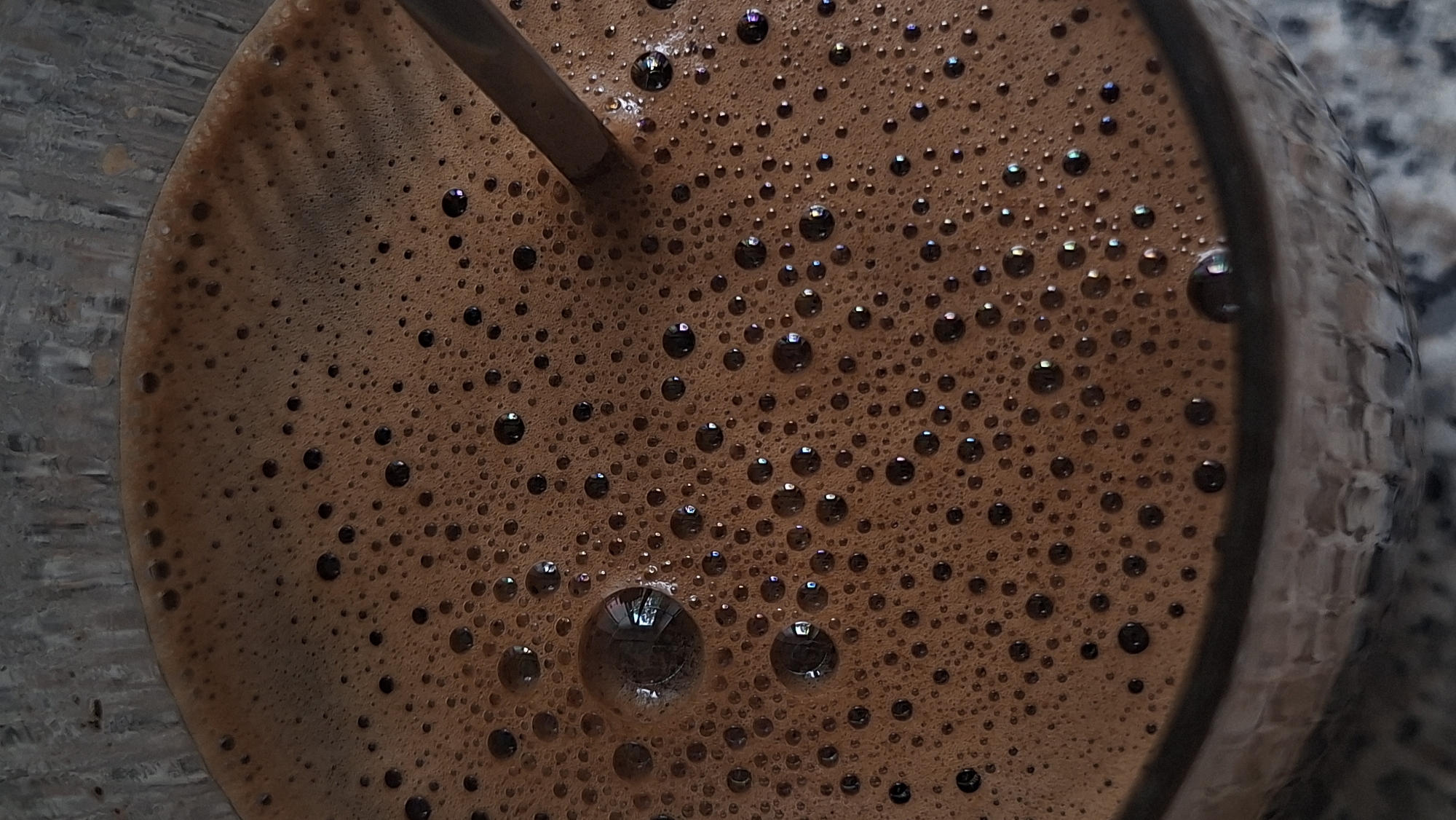 A close-up shot of coffee with a thick layer of foam and bubbles. A metal spoon is partially submerged in the coffee. The coffee is in a clear glass with a textured surface. The background appears to be a grey countertop with some speckling. The lighting is soft, highlighting the foam and reflections on the bubbles. There are no people or discernible city landmarks visible.