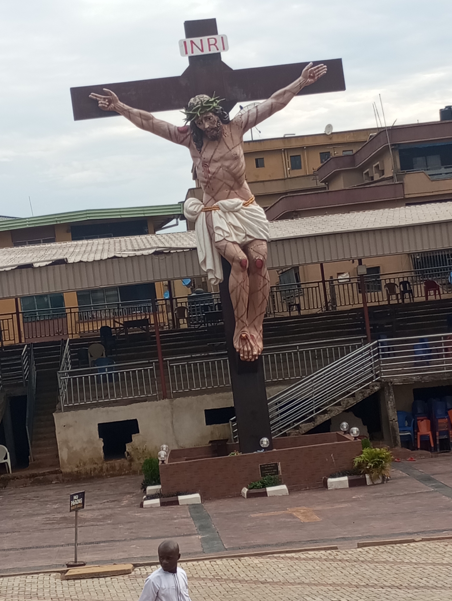 The image shows a large statue of Jesus Christ on the cross, with the inscription "INRI" at the top of the cross. The statue is located in front of a building with multiple stories and balconies, possibly in a public space or a religious institution. There are also stairs and railings visible in the background. In the foreground, a man in a white shirt is walking away from the camera, and a "NO PARKING" sign is posted on a metal pole. The ground is paved with interlocking bricks and concrete. The overall scene appears to be outdoors, with an overcast sky. Based on the available information, this statue is located at the Holy Cross Cathedral in Onitsha, Nigeria.