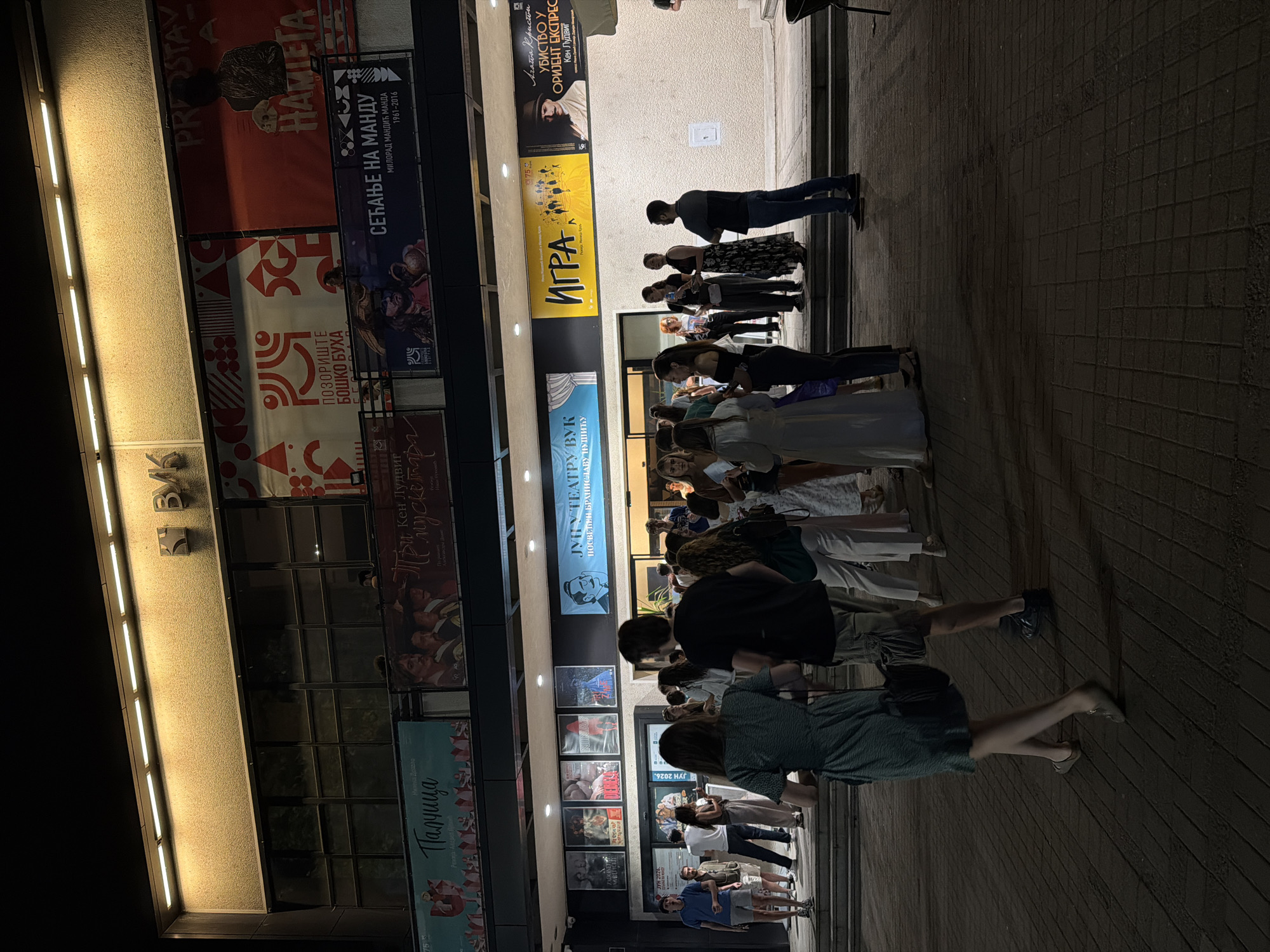 The image shows the exterior of the Vuk Theatre, located in Zvezdara Urban Municipality, Serbia. A crowd of people is gathered outside the theatre entrance at night, appearing to be waiting or socializing. The theatre facade is illuminated by spotlights and features several large posters advertising upcoming performances. People are dressed in casual and semi-formal attire. The ground is paved with brickwork.