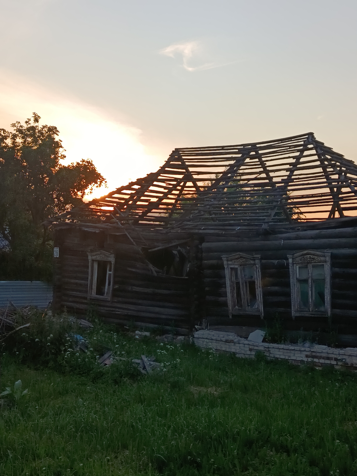 The image depicts the exterior of a dilapidated wooden house at sunset in Motyakovo, Russia. The house, constructed of dark logs, has a severely damaged roof, with the wooden rafters exposed to the sky. The house has two windows visible, both with wooden frames and glass panes that appear to be intact, though weathered. A small sign with the number "26" is affixed to the exterior wall to the left of the left window. The foreground is covered in green grass, with some scattered white wildflowers. To the left of the house, there is a metal fence and piles of debris, including wood and what appears to be construction materials. Behind the house, a large tree with dark green foliage is visible, and the setting sun casts an orange glow through its branches and illuminates the sky with warm tones, with a faint cloud formation visible. The overall scene suggests neglect and disrepair of the structure.
