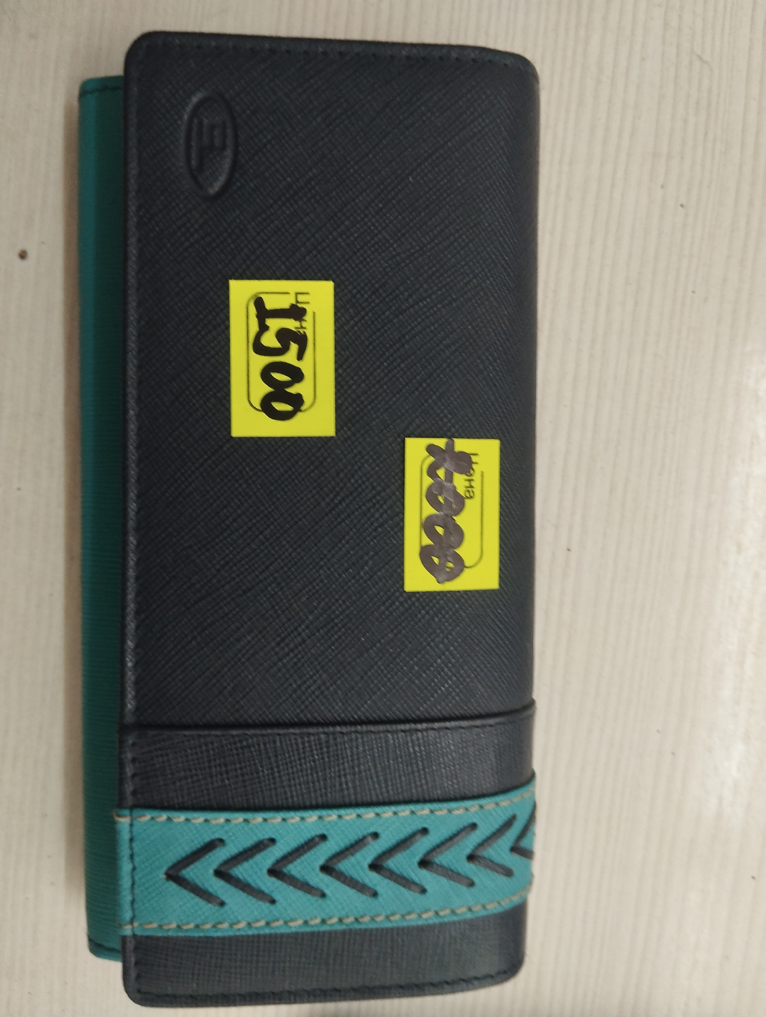 The image shows a black wallet with a teal and black patterned band near the bottom. Two yellow price tags are affixed to the wallet. The tag on the left has "1500" written in black marker. The tag on the right has "800" written in black marker with "BHC" below it. The wallet is lying flat on a light-colored, wood-grained surface. The image appears to be taken indoors, with no discernible features of the city of Tokmok, Kyrgyzstan.