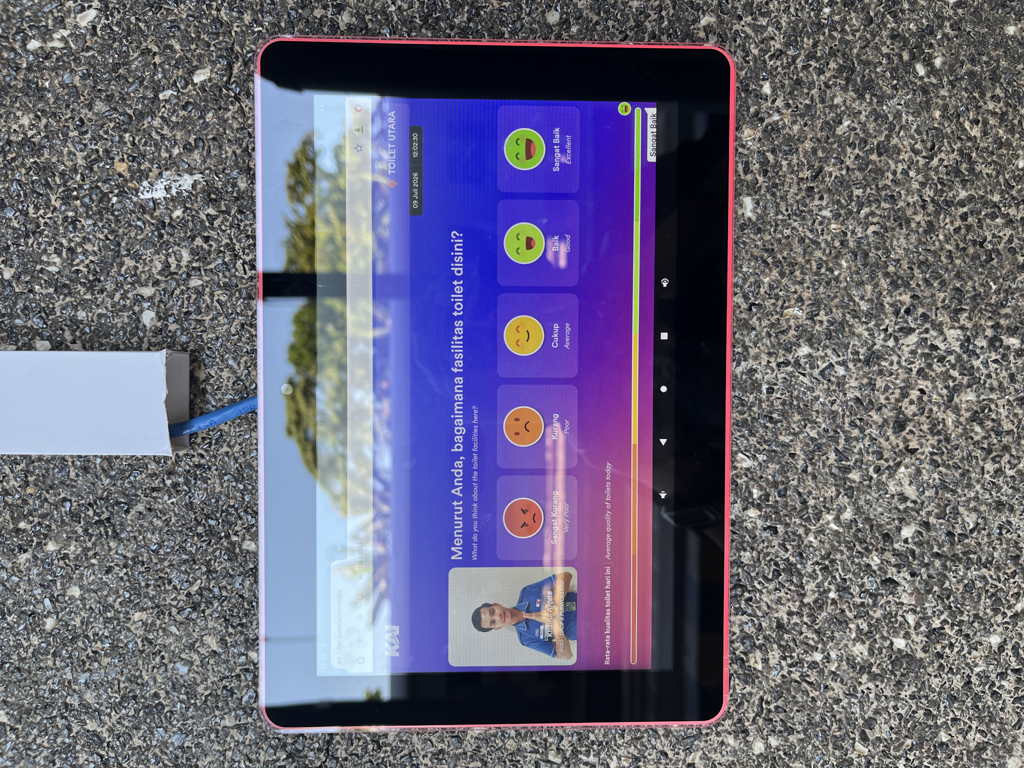 An outdoor eye-level medium shot shows a tablet computer mounted on a textured, gray, pebbled surface, likely a wall or sidewalk, in Desa Tambakreja, Indonesia. The tablet, with a bright pink casing, displays a digital survey about toilet facilities. The screen shows a photograph of a young man in a blue uniform, identified as Khairul Anjal, along with a question in Indonesian and English: "Menurut Anda, bagaimana fasilitas toilet disini? What do you think about the toilet facilities here?". Below the question are five emoji-based rating options: "Sangat Kurang" (Very Poor), "Kurang" (Poor), "Cukup" (Average), "Baik" (Good), and "Sangat Baik" (Excellent). A progress bar at the bottom of the screen indicates "Rata-rata kualitas toilet hari ini | Average quality of toilets today," with the "Sangat Baik" option highlighted. Above the survey, a date and time are visible: "09 Juli 2026 12:02:30" and "TOILET UTARA." A white electrical box with a blue cable exiting is attached to the pebbled surface above and to the left of the tablet. The background is blurred, showing green foliage and a bright sky, suggesting an outdoor, daytime setting.