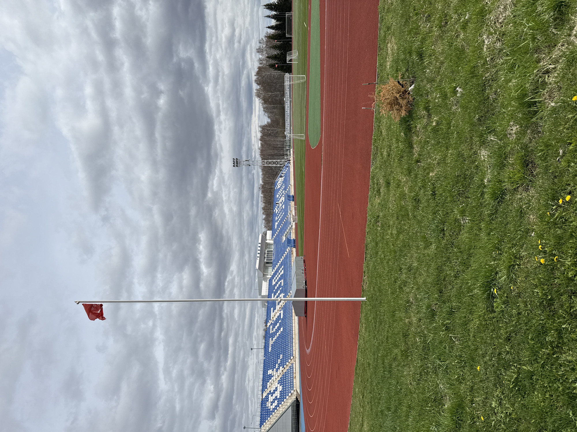 The image depicts an outdoor scene at a stadium or sports complex in Tobolsk, Russia.
The scene includes a red running track, an adjacent grassy field, and stadium seating with blue and white seats. There is a flag pole with a red flag at the side of the stadium.
In the background are a soccer field with goalposts and trees. The sky is overcast with gray clouds. The scene is devoid of human subjects or visible actions.