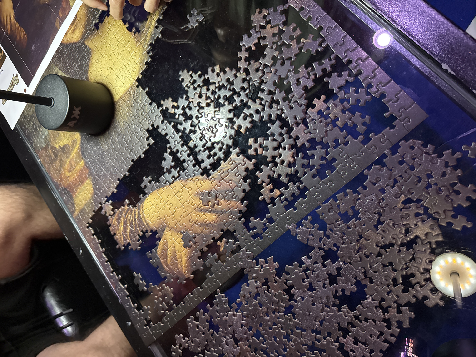 The image shows a partially completed jigsaw puzzle on a dark, reflective surface, likely a table. The puzzle pieces are metallic gray and appear to be made of metal. The puzzle image itself is visible in the upper right portion of the frame, featuring a golden, textured design that appears to be an artistic rendition of fireworks or a similar explosive visual. A black, cylindrical object with white text that reads "АЖ" is positioned on the puzzle, likely a lamp base or a piece of equipment. In the lower left and right corners, there are illuminated LED lights, one white and one purple, which appear to be part of the table or surrounding environment. In the background, parts of a person's legs and torso are visible, seated in a black chair. The overall scene suggests a recreational activity or a workspace. The image does not contain enough information to determine its location within Phuket City Municipality, Thailand.