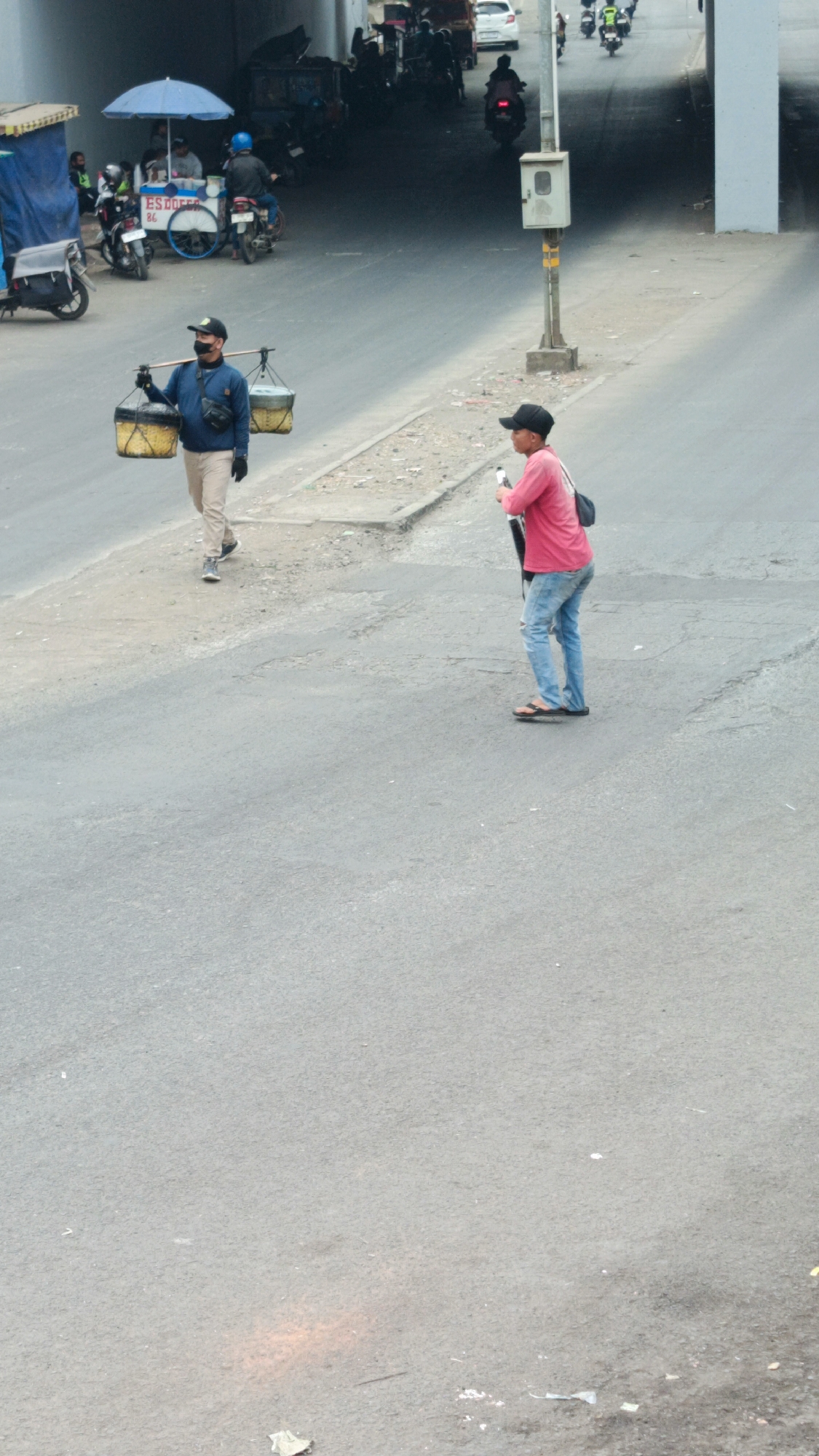 In Margajaya, Indonesia, two individuals are walking on a paved road. The person on the left, wearing a blue long-sleeved shirt, light-colored pants, and a black baseball cap, carries two cylindrical baskets suspended from a wooden pole across their shoulders. A dark bag is slung across their chest. The person on the right, dressed in a bright pink long-sleeved shirt, ripped blue jeans, and a black baseball cap, holds a long, dark object vertically. This individual is wearing dark flip-flops. The road appears to be an urban environment with a pedestrian walkway to the left, which has various stalls and parked motorcycles. In the background, a concrete overpass casts a shadow over a section of the road, and several motorcycles are visible moving under it. The overall impression is a bustling street scene with individuals engaged in their daily activities.