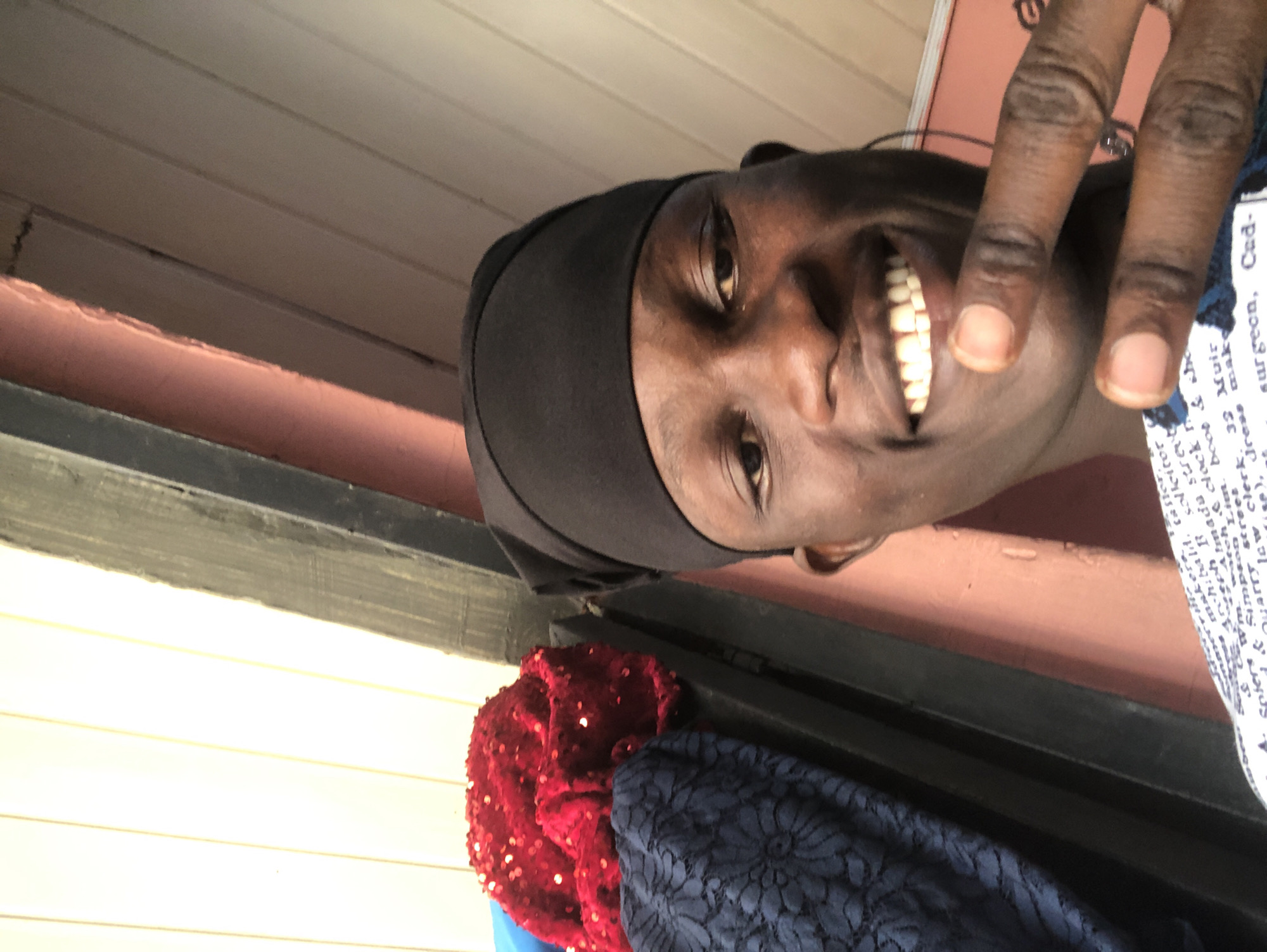 A close-up, self-taken photograph shows a young Black man wearing a black durag. He is smiling and holding up two fingers in a peace sign. The man is in the foreground, with the background showing a pink wall and wooden paneling. To the left of the man, there are stacks of fabric, one red and sequined, the other dark blue and patterned. The lighting is bright, casting shadows on the man's face. The image appears to have been taken indoors, possibly in a residential setting. The presence of fabric stacks suggests the man may be involved in tailoring or seamstress work, or that he is in a place where such materials are stored. The overall impression is a casual, informal portrait.