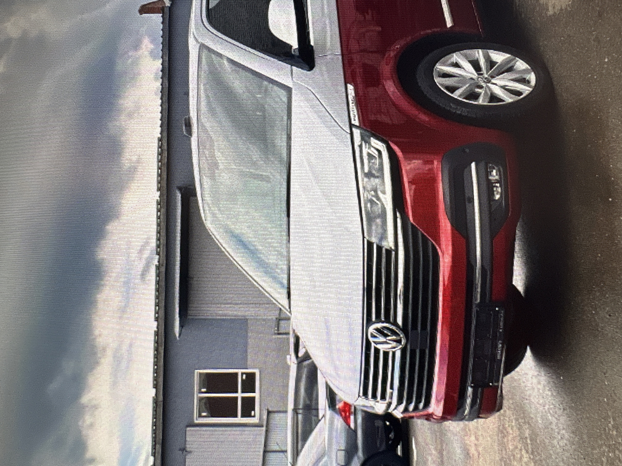 A red and silver Volkswagen Transporter van is parked on a wet asphalt surface in front of a grey industrial building with a large window. The sky is overcast with dramatic clouds. The van's front grille features the Volkswagen logo, and the license plate is visible. Another dark-colored car is parked to the left of the van, partially obscured. The scene appears to be an outdoor storage or parking area.