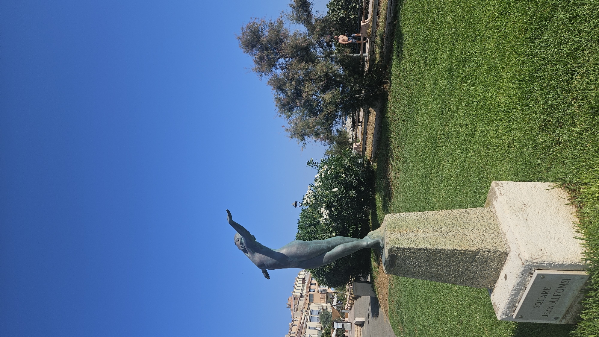 The image depicts a bronze statue of a female figure in a dynamic pose, with her arms outstretched upwards and her body arched backwards. The statue is positioned on a stone pedestal in a well-maintained grassy area. A plaque at the base of the pedestal reads "SQUARE JEAN ALFOnSI". The background reveals a clear blue sky, buildings with terracotta roofs, lush green bushes, and a large tree. To the right, a man wearing swim shorts walks along a path. The scene appears to be a public square or park in a coastal town, likely L'Île-Rousse, France, given the inscription on the plaque.