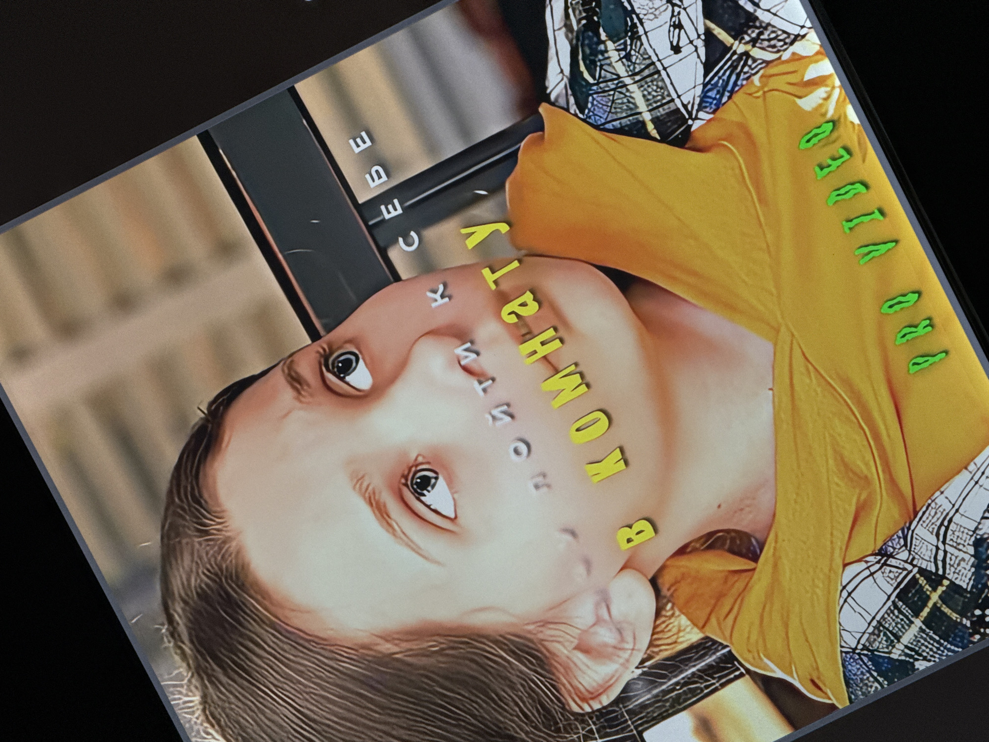 The image is a close-up, angled photograph of a young person's face. The person has light-colored skin and dark hair. They are wearing a bright yellow t-shirt and a black and white plaid shirt underneath. The image is overlaid with text. On the person's forehead, yellow letters spell out "В КОМНАТУ" (V KOMNATU), which translates to "in the room" in Russian. Below that, on the person's cheek, are smaller, lighter colored letters that appear to spell "К" and "И". To the left of the face, on a dark object that appears to be a shelf or frame, white letters spell out "СЕБЕ" (SEBE), meaning "for oneself" in Russian. On the sleeve of the yellow t-shirt, green letters spell out "PRO VIDEO". The lighting suggests an indoor setting, possibly a studio or home, and there are no clear indicators of time of day or weather. The overall emotion conveyed is ambiguous due to the stylized text and angle of the photograph. The background is blurred, with hints of what could be bookshelves or a similar interior setting.