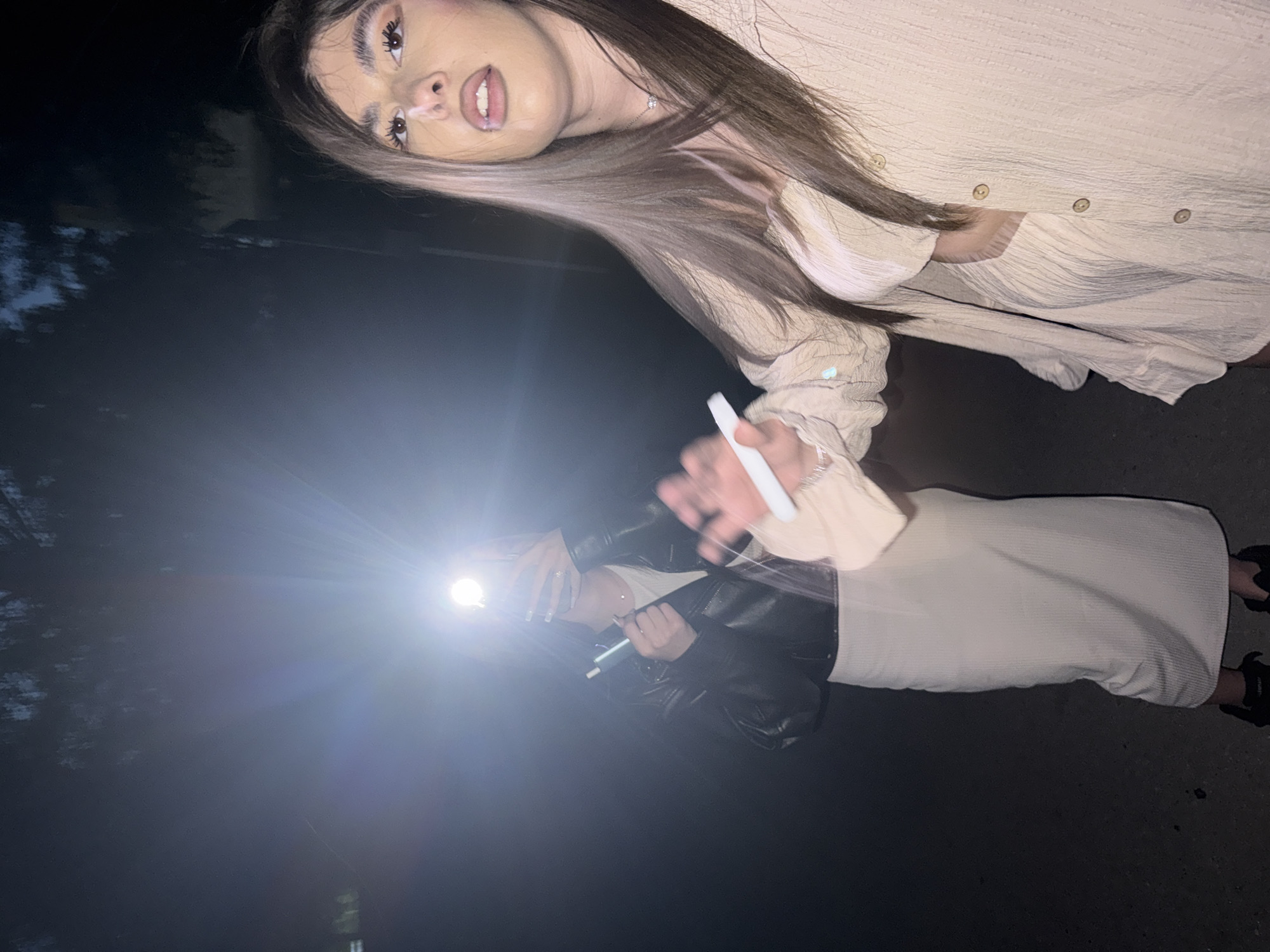 The image depicts an outdoor, nighttime scene within the city of Blagoevgrad, Bulgaria, illuminated by a strong, artificial light source. The ground is a dark, possibly paved surface, and the background shows dark silhouettes of trees against a night sky.
Two females are present. In the foreground, positioned on the right, a female with long, multi-toned brown hair wears a light-colored, textured two-piece outfit consisting of an open long-sleeved button-up shirt over a lighter top. Her face, partially visible, displays prominent eye makeup and a white substance on the bridge of her nose; her mouth is slightly open. Her right arm is extended, holding a white cylindrical object. She also wears a thin silver necklace.
In the mid-ground, on the left, a second female is partially obscured by intense light originating from a smartphone held in her right hand, pointed towards the camera with its flashlight activated. She is wearing a dark, possibly leather, jacket over a white top and a long, light-colored ribbed skirt. Her left hand holds a teal-colored cylindrical object.
The scene is characterized by dramatic, high-contrast lighting from the direct flashlight, resulting in lens flare and strong shadows, with the ambient environment remaining very dark. Both individuals are holding objects consistent with electronic vaping devices.