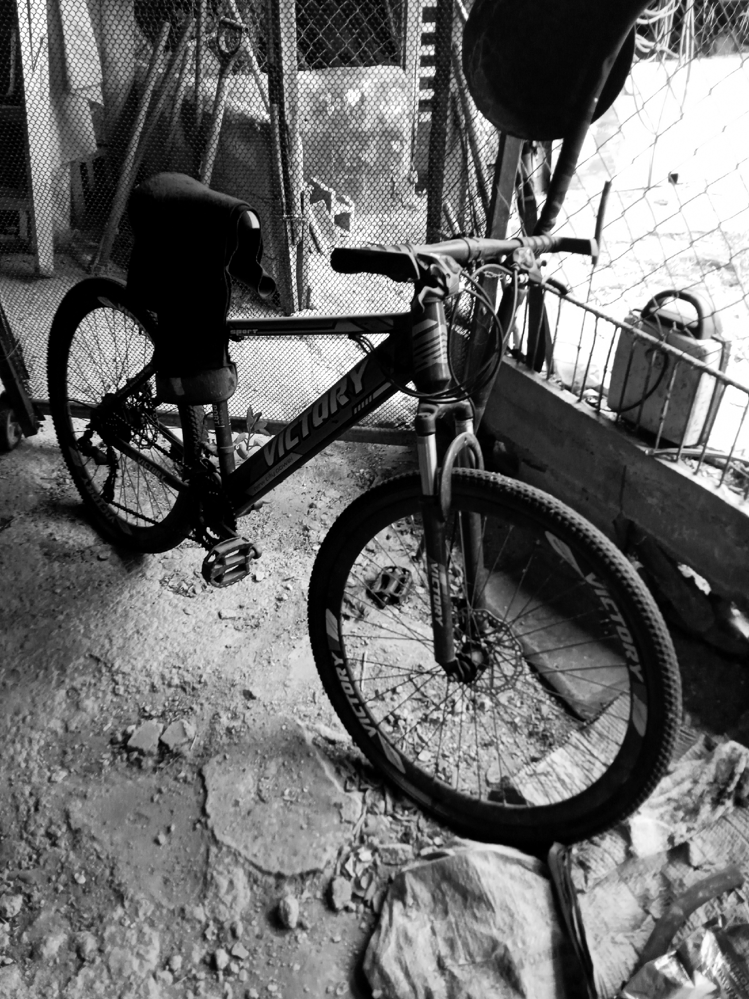The image depicts a bicycle, brand "VICTORY", parked in a cluttered indoor space. The location appears to be a storage area or shed, possibly in Montalbán, Venezuela. The background includes a chain-link fence and various indistinct objects. The ground is a mix of dirt and debris. No people are visible.