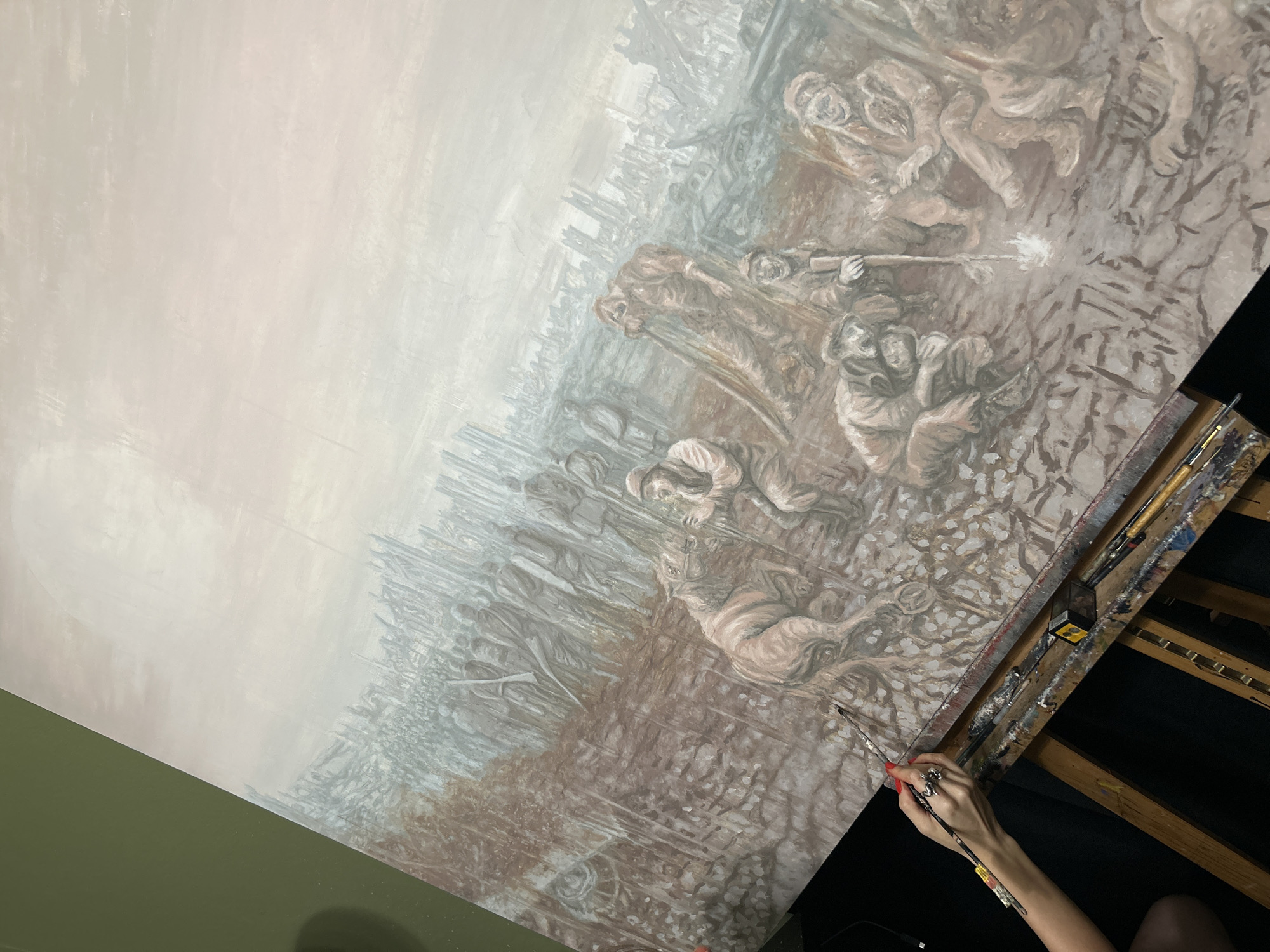 A work-in-progress painting depicts a war scene with figures engaged in combat. Some figures are armed with rifles, one of which is depicted firing. The style is reminiscent of historical war art. The painting is situated on an easel, alongside an array of paintbrushes. It is not possible to determine any location as being Cape Coral, United States of America.
