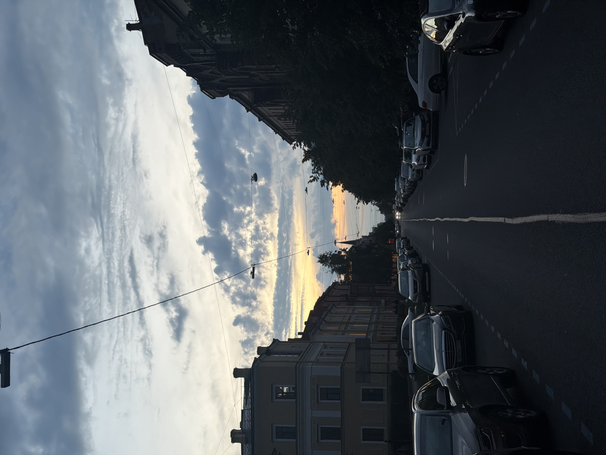 The image depicts a street in Saint Petersburg, Russia, at dusk. The sky is filled with dramatic clouds tinged with the warm hues of the setting sun. Buildings line both sides of the street, with a lighter-colored building on the left and a darker, more imposing structure on the right, partially obscured by trees. Numerous cars are parked along the street, both on the left and right sides, and some are visible on the road. The street itself is paved, with visible lane markings. Overhead, power lines crisscross the sky, with lampposts visible. The overall atmosphere is that of a quiet urban evening.
