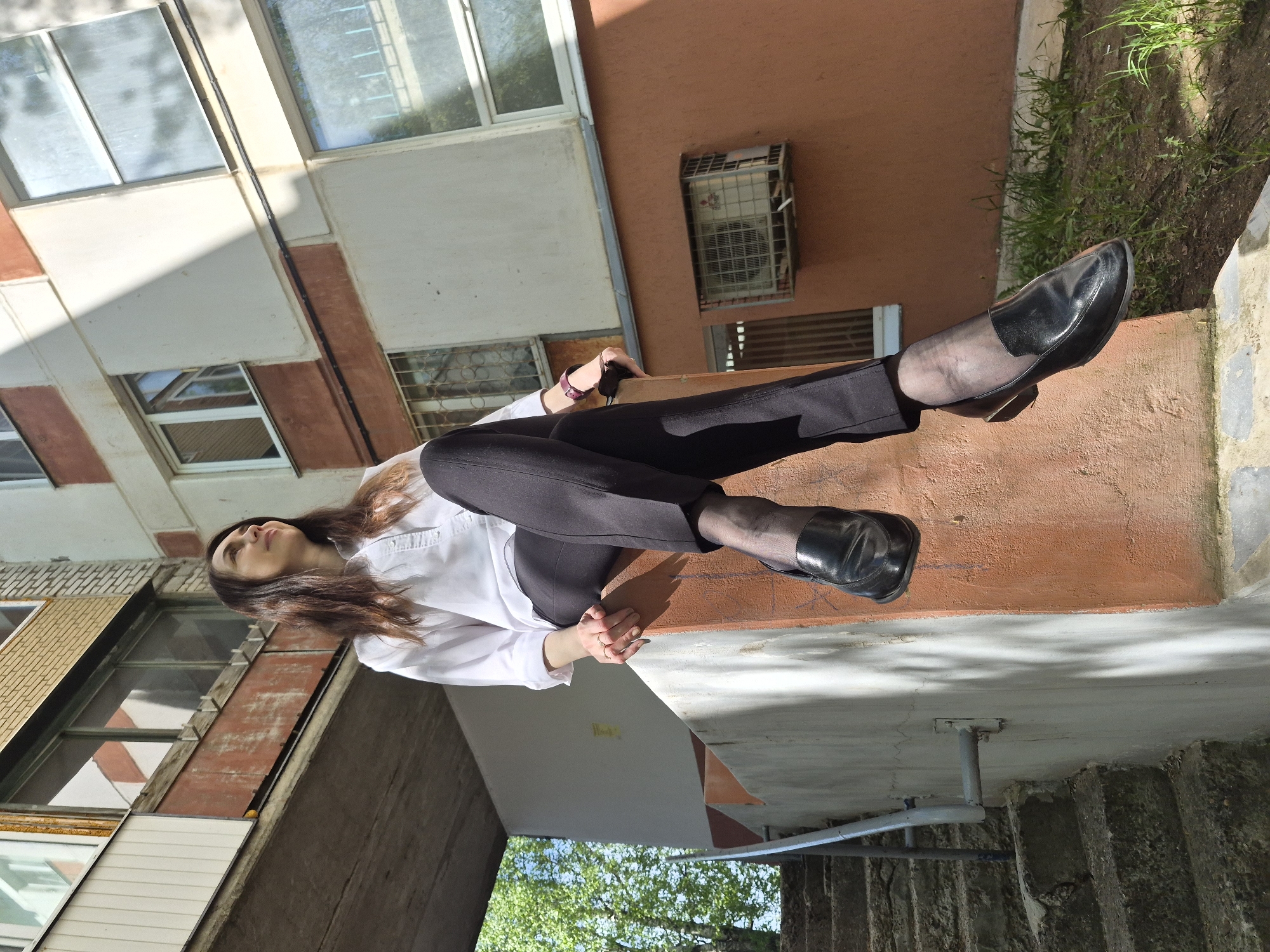 A woman is seated with her legs stretched out in front of her. She is wearing black pants, a white shirt, black stockings, and black shoes. She is sitting on a ledge that appears to be outside a building. The building has a red wall and white window frames. There is some grass and a tree visible in the background. The location appears to be an urban area with a mix of buildings and greenery.