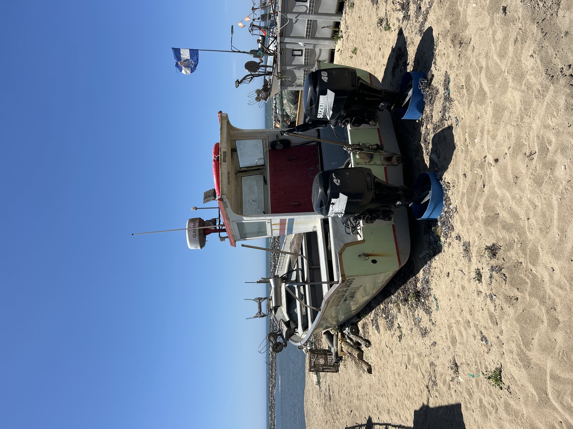 A long, narrow fishing boat with a white cabin sits on a sandy beach under a clear blue sky. The boat has two outboard motors, both bearing the Suzuki logo and the number 40. A Garmin radar dome is mounted on the cabin roof. A blue and white flag, possibly a provincial flag, is attached to a flagpole at the rear of the boat. The boat is beached above the waterline, with the ocean and a breakwater visible in the background. Several other fishing vessels and equipment are present on the beach, suggesting a working harbor or fishing village. The sand is coarse and scattered with debris.
