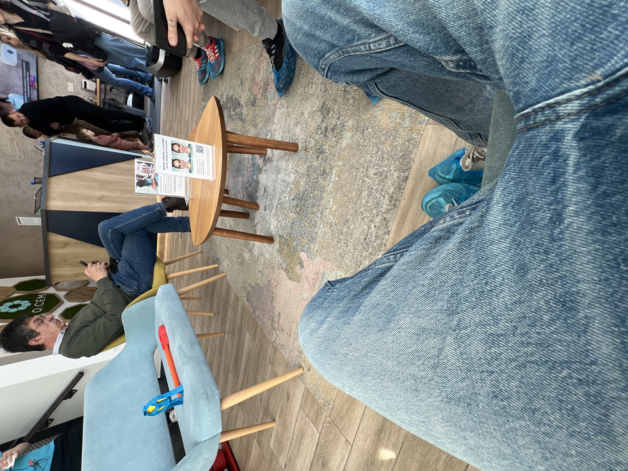 The image depicts an interior waiting area in Burgas, Bulgaria, possibly in the OSEM business building, suggested by the O.CEM logo visible on the wall. The scene focuses on waiting individuals, with emphasis on their lower bodies and legs. Several people are wearing blue disposable shoe covers. Jeans are a common garment, with the close-up of denim dominating the view. A small round table stands near several chairs, on which sits a printed informational flyer, presumably for the OSEM business. A man is seen on a light blue chair with children's toys sitting to the right side of him. Other people are standing nearby, and there is a blue counter or desk in the background of the scene.