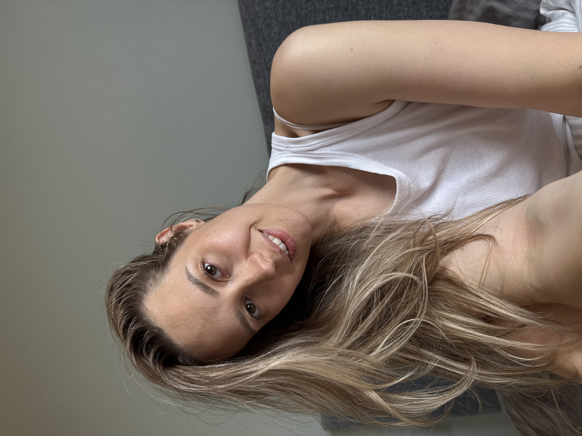 A woman with long, light brown hair and brown eyes is lying down, facing the camera with her head tilted. She is wearing a white tank top. The background consists of a gray wall and part of a gray sofa. The image does not contain enough information to determine a location in Chaikovsky, Russia.