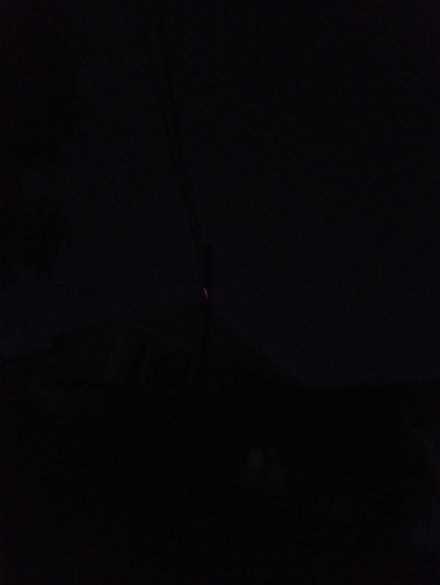 The image appears to be taken at night in Jos, Nigeria. The scene is mostly dark, with the vague outlines of rooftops visible against the dim sky. A thin vertical element, possibly a wire or pole, is subtly illuminated with a tiny, faint reddish light at its middle point. The rest of the foreground is consumed by shadows, lacking distinguishable details. The overall environment seems quiet, with no apparent activity or presence of people.