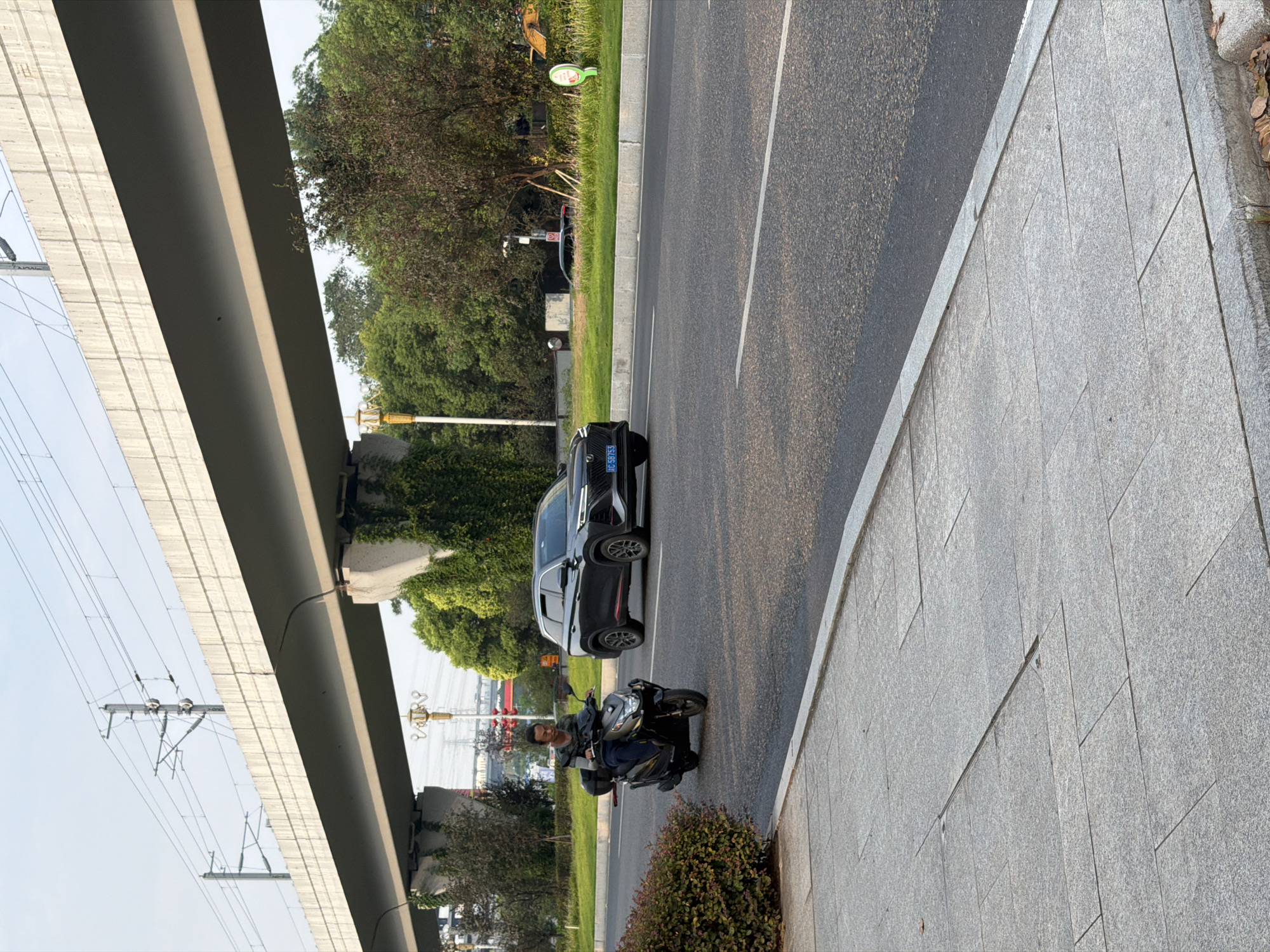 An eye-level, medium shot shows a person riding a scooter on a road under a concrete overpass, likely in Jiangjin, China. The person, wearing a camouflage-patterned jacket and dark pants, is looking forward. The scooter, a dark grey model with a storage box on the back, is moving from left to right. To the right of the scooter, a black SUV is also traveling in the same direction. The SUV has a blue license plate with white characters. The road surface is asphalt, with a white dividing line. To the left of the road, a paved sidewalk runs parallel, bordered by a manicured hedge. The overpass, constructed of concrete, spans across the upper portion of the image, with overhead power lines and poles visible. Green trees and foliage are present behind the road and under the overpass. The sky is clear with a light haze.