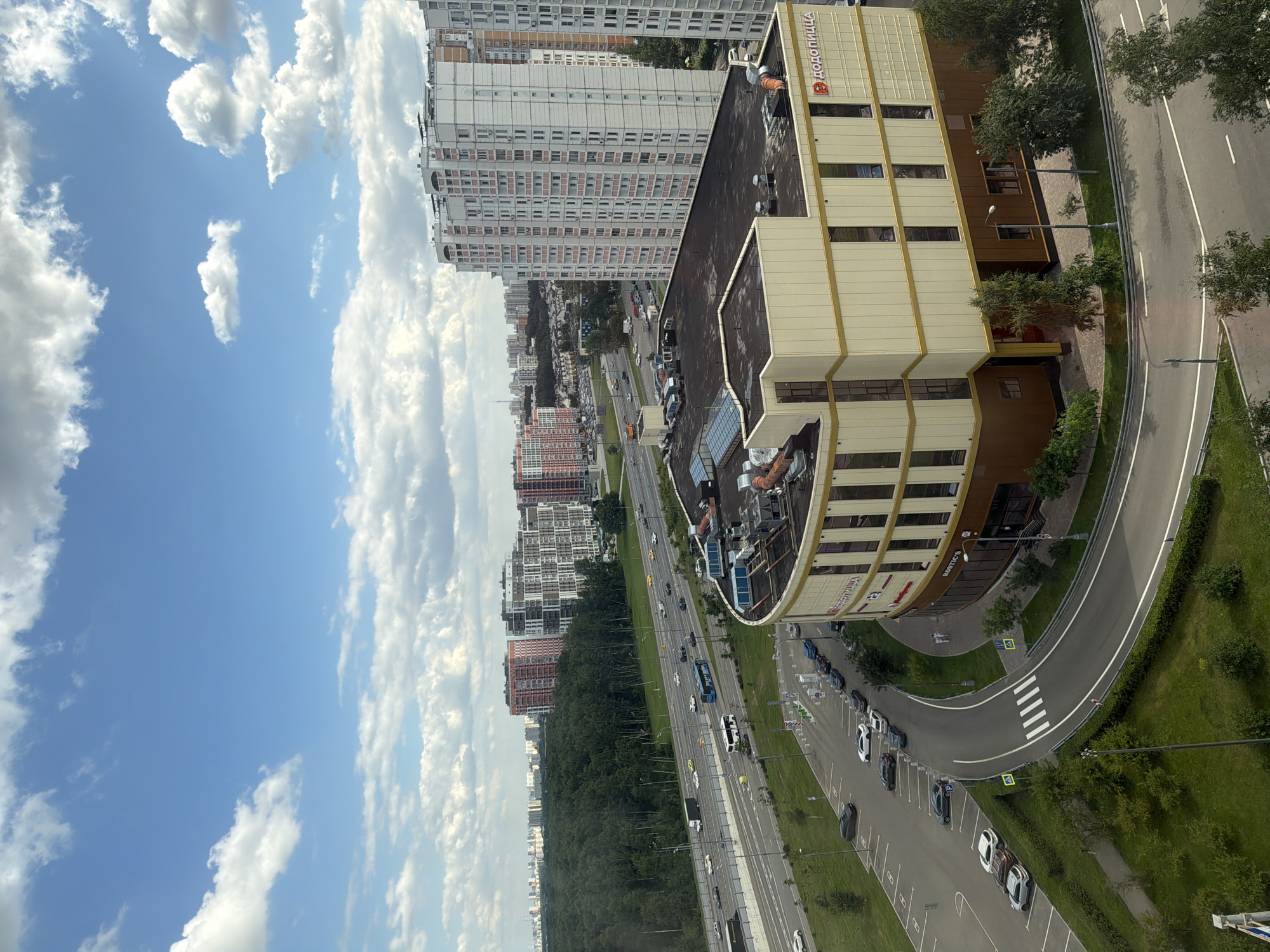 An eye-level, medium shot captures a commercial building with a beige and brown facade, featuring a large sign with Cyrillic lettering reading "Додо Пицца" on its upper section. The building is situated in an urban environment, with a multi-lane highway visible to its left, carrying various vehicles including cars and a blue bus. A parking lot with several cars is located in front of the building. In the background, a dense forest area is present, beyond which numerous high-rise residential buildings and some red-accented apartment complexes are scattered across the cityscape under a bright, cloudy sky.
