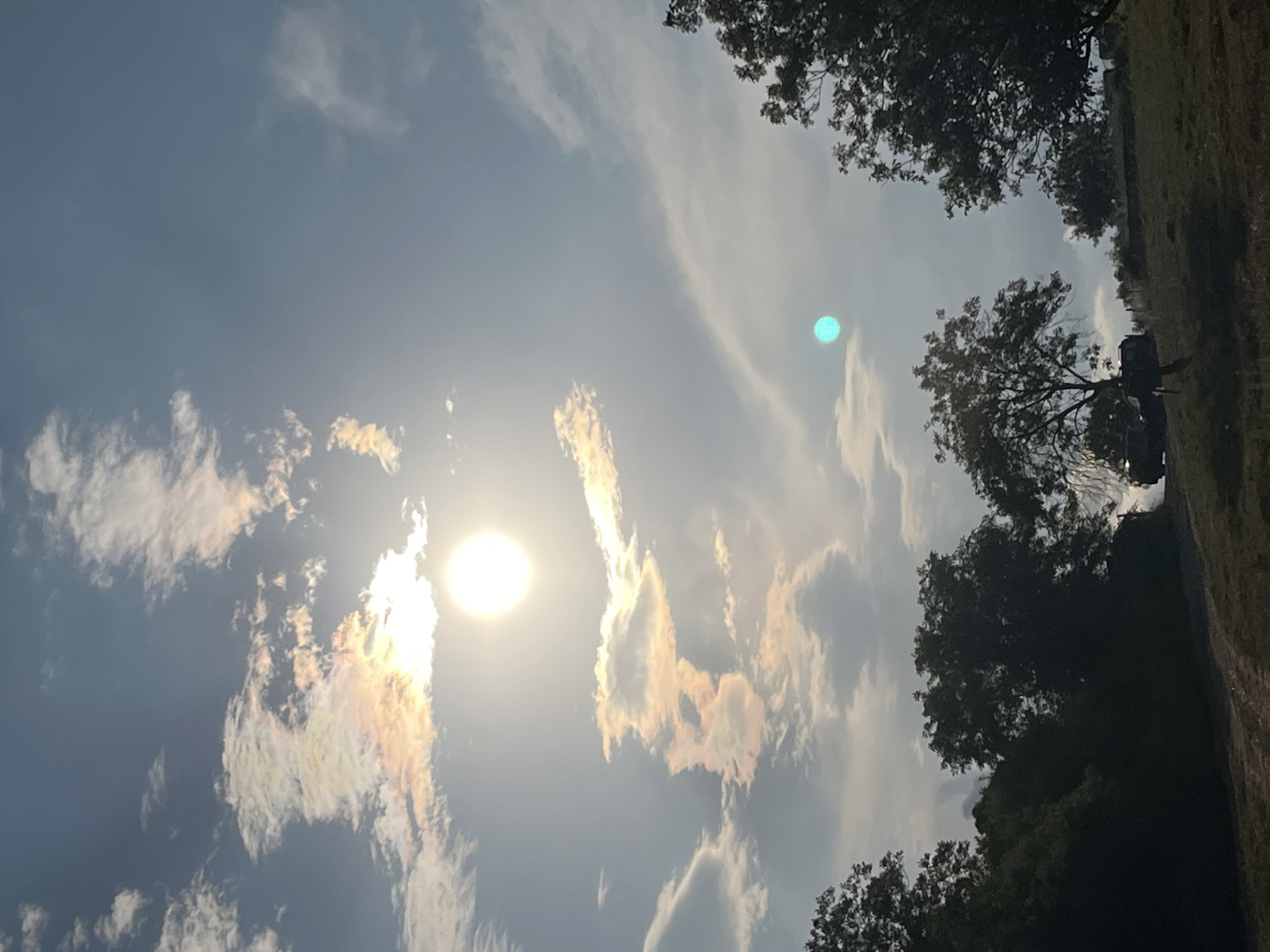 The image captures a bright, partially obscured sun in a hazy sky, with iridescent clouds reflecting pastel hues of pink, yellow, and blue. Below the sky, silhouetted trees with full foliage line a sloping hill. Two dark-colored vehicles are partially visible amongst the trees on the hilltop. A dirt path winds through the foreground. The scene suggests an outdoor environment, possibly a rural or park area, given the vegetation and the presence of vehicles. The lighting indicates it is daytime, with the sun's intensity softened by atmospheric conditions. The location in Ahtopol, Bulgaria, implies a coastal or near-coastal setting, which could contribute to the hazy sky.